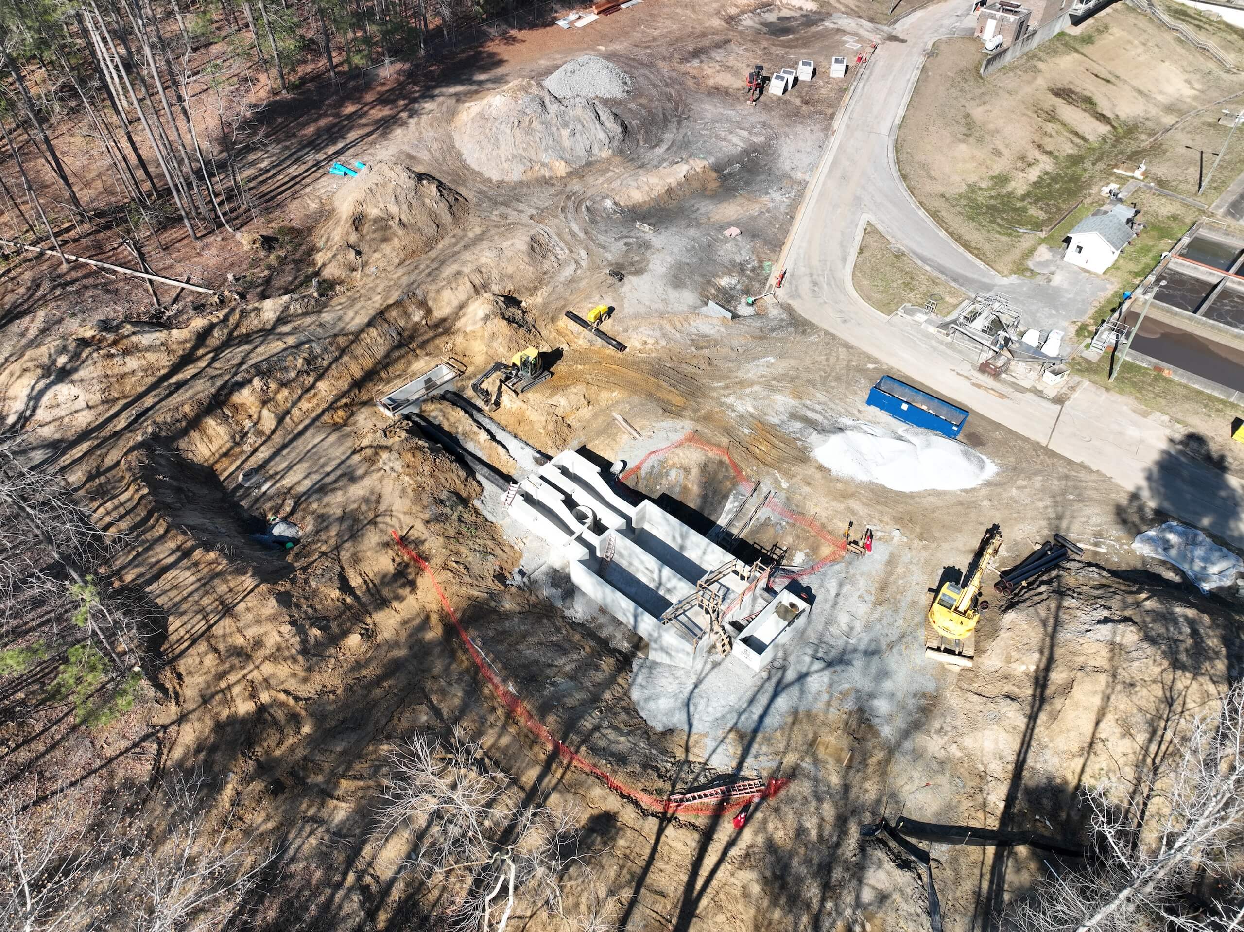 Aerial view of a construction site with heavy machinery, dirt piles, and a partially built concrete structure surrounded by trees and equipment. Red fencing marks the work area.