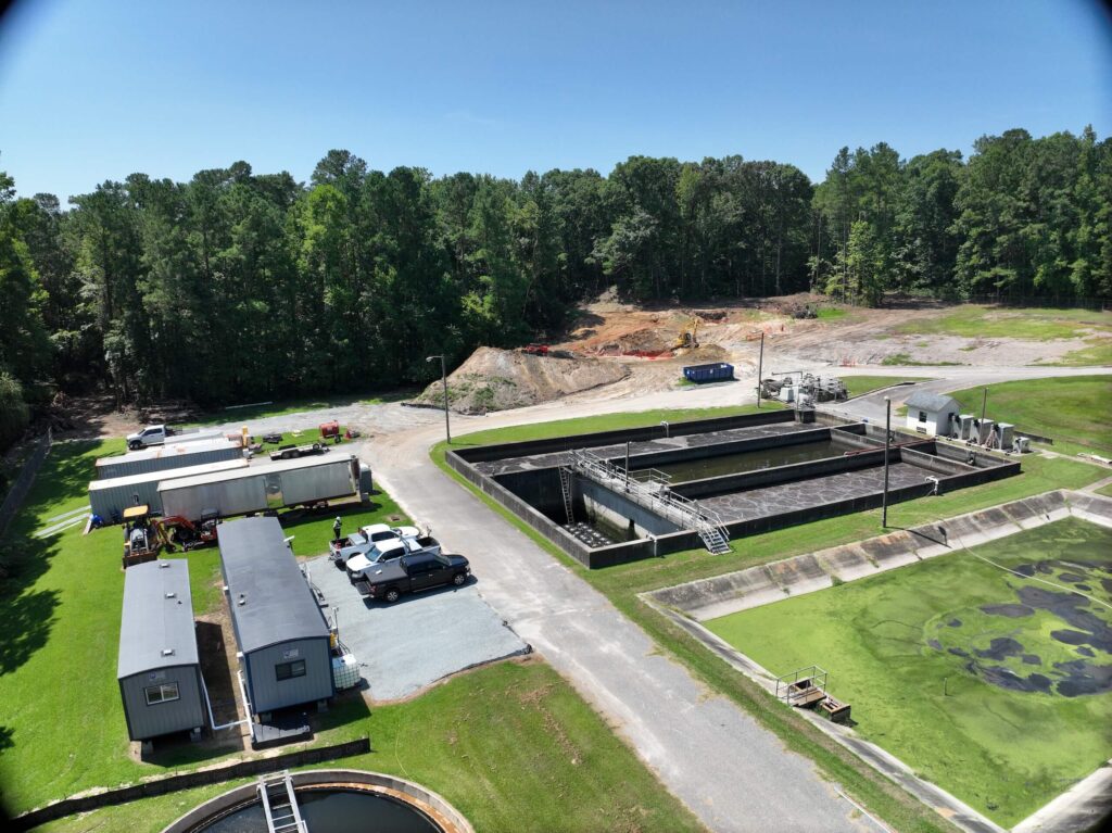 Aerial view of a wastewater treatment plant surrounded by trees, with rectangular treatment basins, parked vehicles, temporary buildings, and construction equipment visible on cleared land in the background.