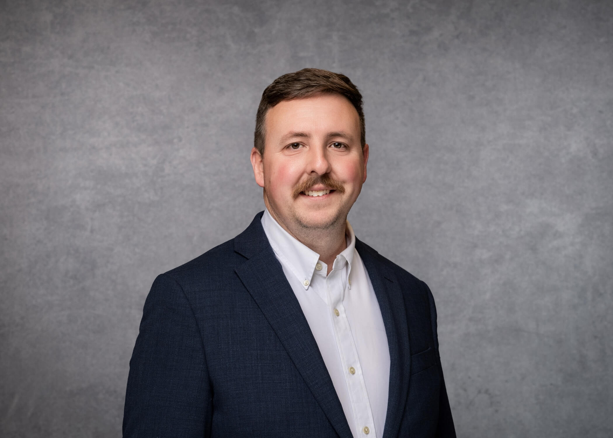 A man with short brown hair, mustache, and light skin, wearing a navy blazer over a white collared shirt, smiles in front of a gray studio background.