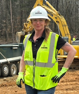 A woman wearing a hard hat, neon yellow safety vest, gloves, and glasses stands on a construction site with a Komatsu excavator and a truck in the background.