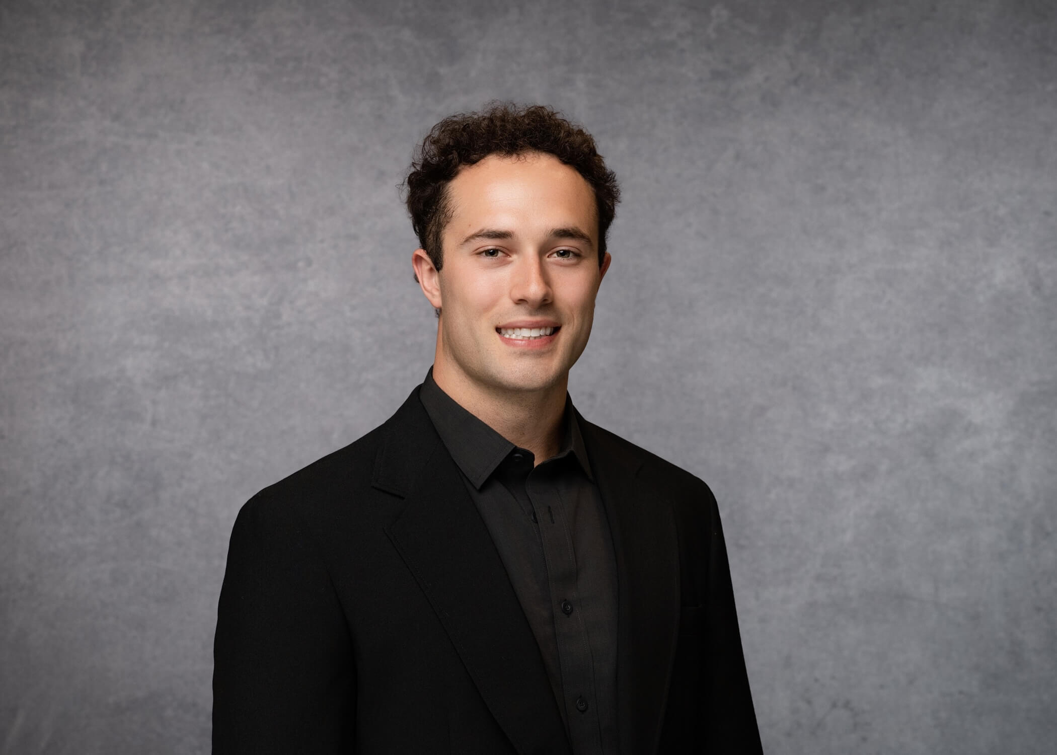 A man with short curly hair wearing a black suit and black shirt stands against a gray textured background, smiling softly at the camera.