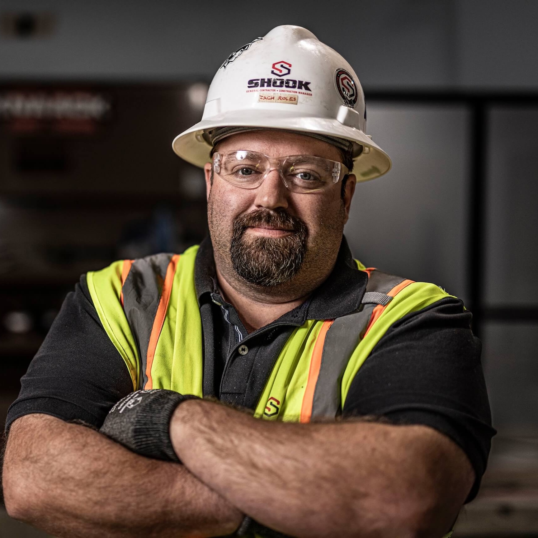 A construction worker with glasses, a helmet, and a neon safety vest stands with arms crossed, looking confidently at the camera.