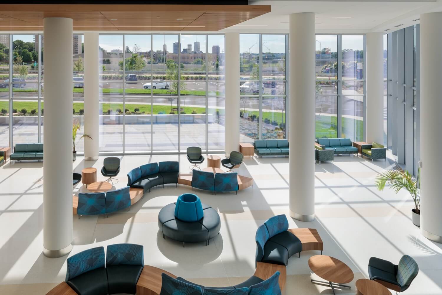 Spacious modern lobby with large windows, round and curved blue seating, wooden tables, and views of a cityscape and green area outside. Natural light fills the room.