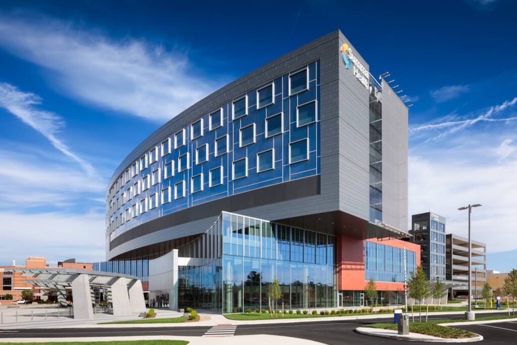 A modern hospital building with large glass windows, a curved facade, and blue sky above. There are landscaped grounds and a parking garage nearby.