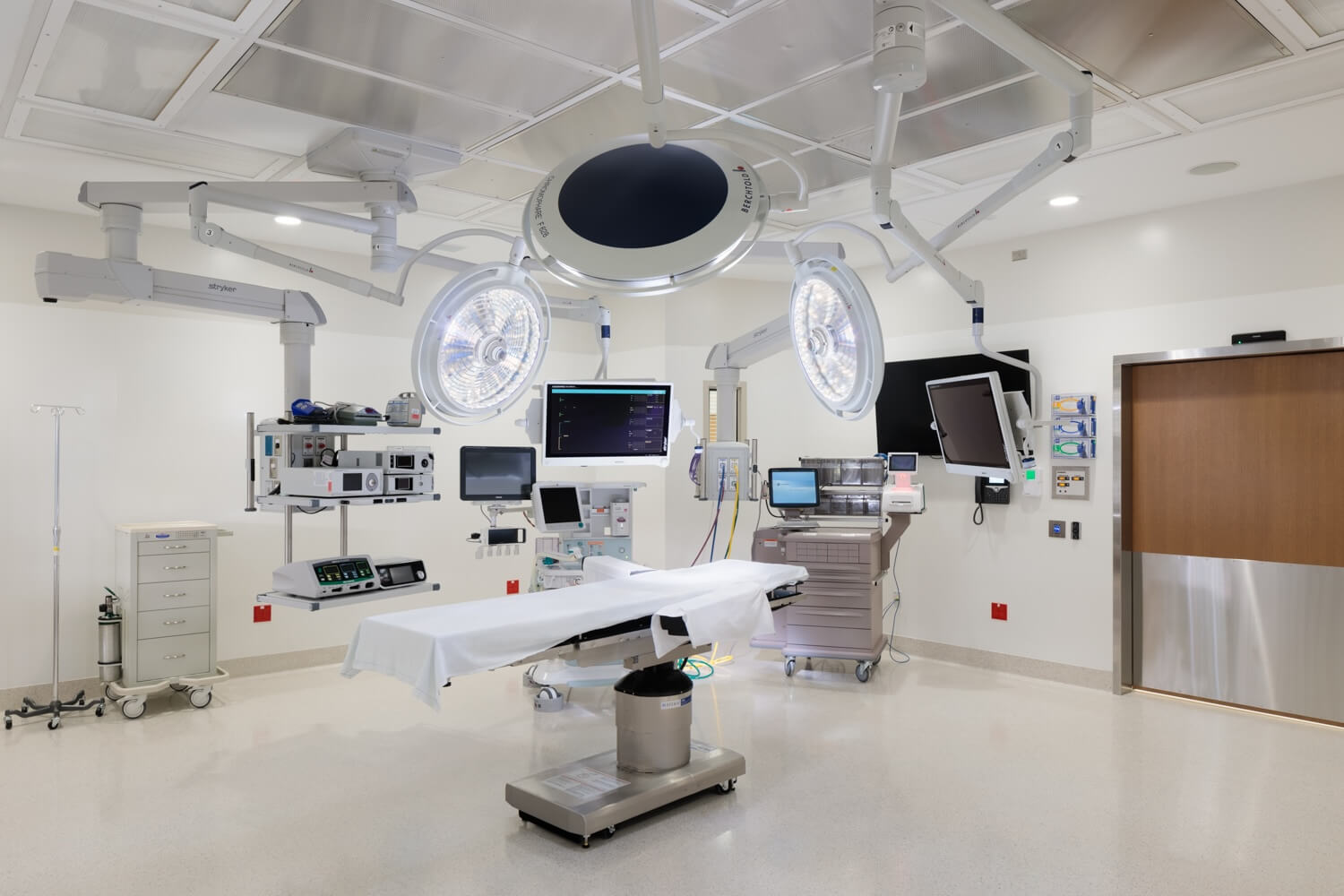 A modern operating room with surgical lights, an empty operating table, medical monitors, equipment carts, and cabinets, all in a clean, sterile environment.