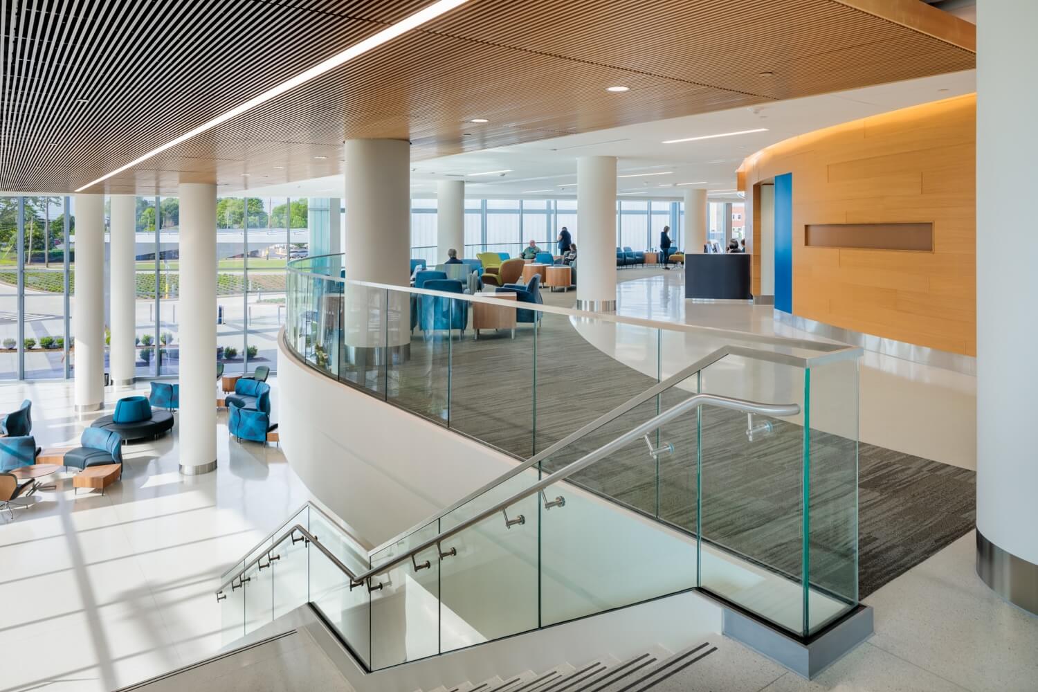 Modern building interior with large windows, glass railings, a staircase, white columns, wood ceiling, and blue chairs arranged in seating areas. Natural light fills the space, creating a bright and open atmosphere.