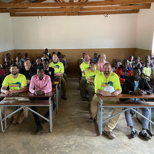 A group of adults, some in bright yellow shirts, sit at desks in a classroom with wooden beams and neutral walls, facing the camera and smiling.