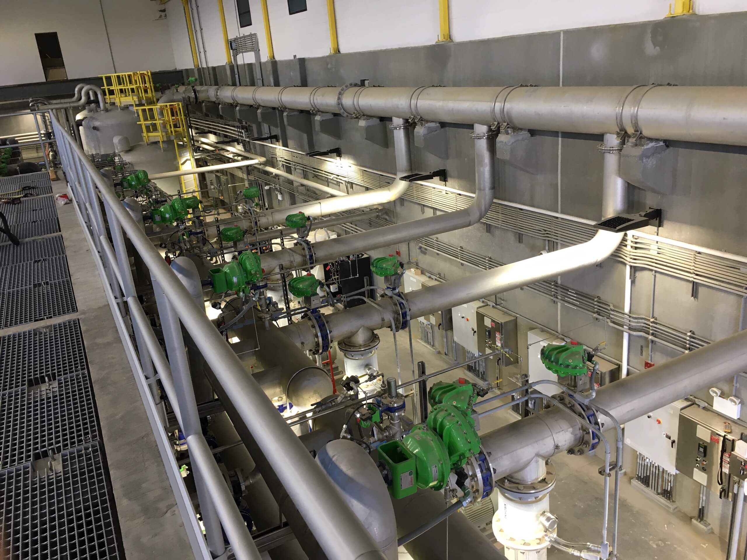 Industrial facility interior with multiple large metal pipes, green valve actuators, and grated floor walkways. Yellow railings and control panels are visible along the walls. The scene is well-lit and orderly.