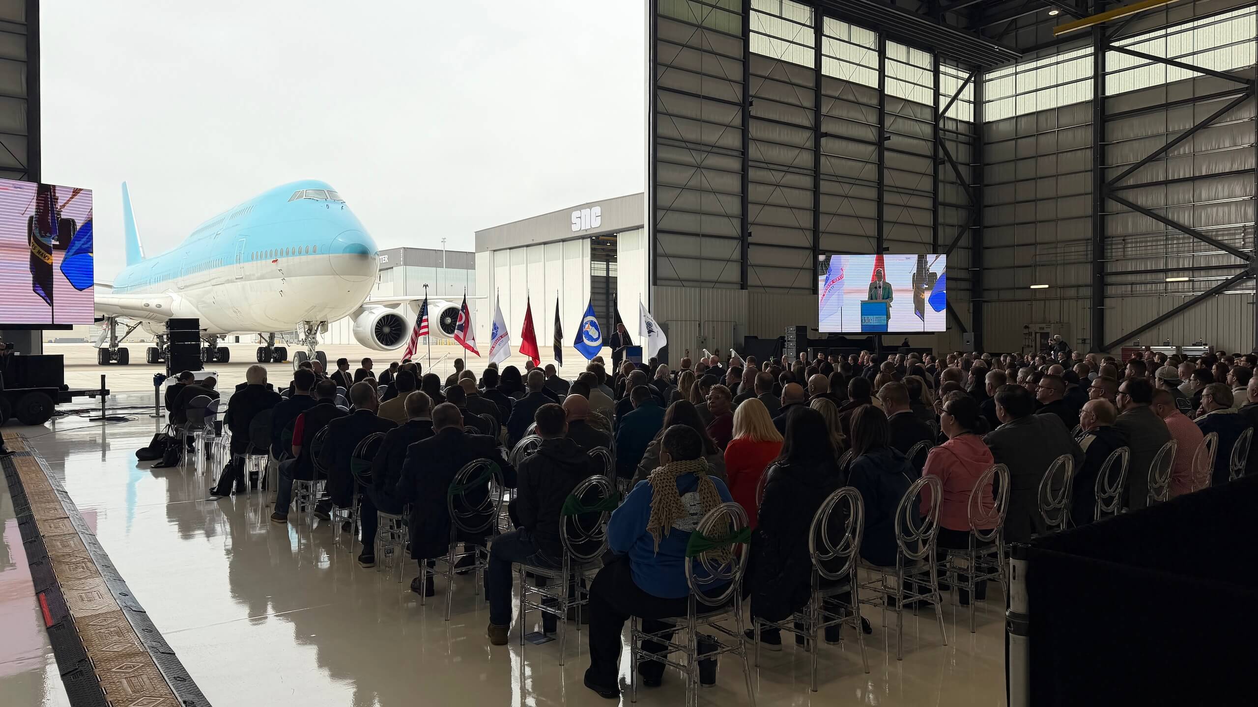 A crowd sits in a hangar facing a large airplane and stage with flags. Two screens display a speaker at a podium. The scene is indoors, with the airplane partially inside the hangar.
