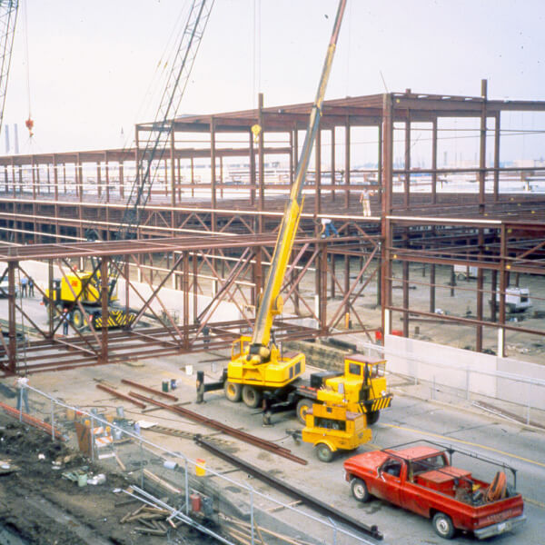 Steel-frame building under construction with cranes, a red pickup truck, construction materials, and workers on site. Machinery and beams are visible around the partially built structure.