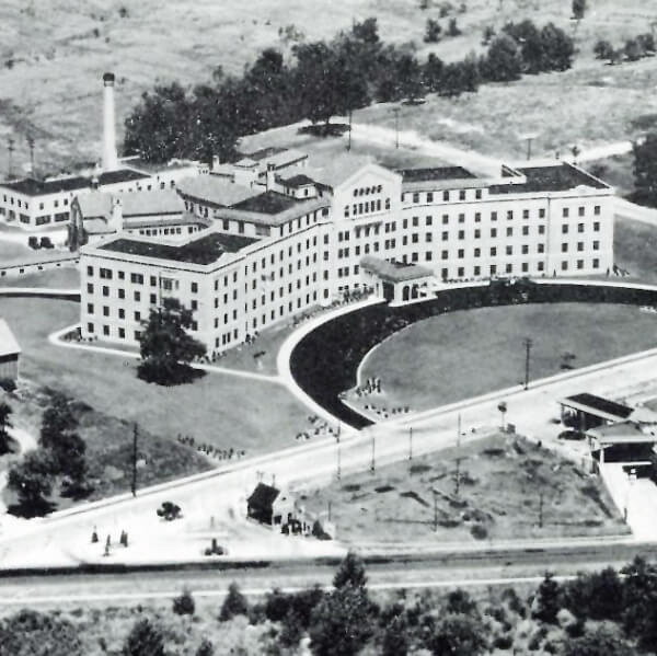 A black and white aerial photo of a large multi-story building with a curved driveway, surrounded by grass, trees, and nearby smaller structures. Roads and train tracks run in front of the property.