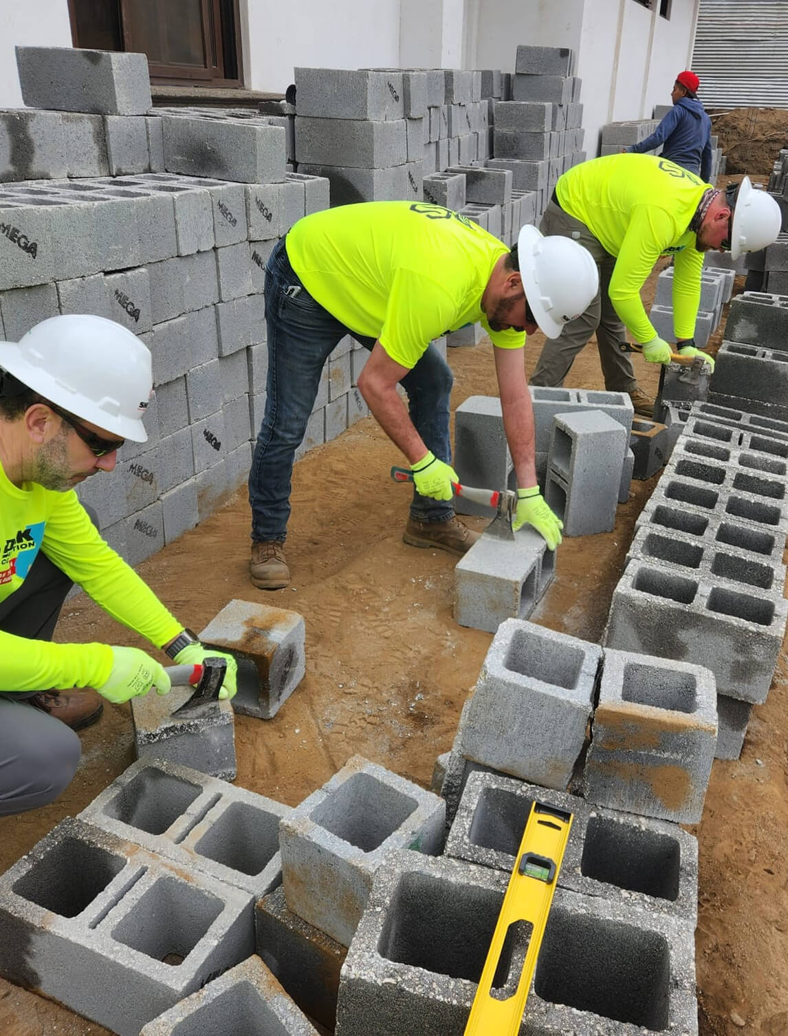 Three construction workers in yellow shirts and white helmets stack and measure concrete blocks at a building site, with stacks of blocks and a level tool on sandy ground.