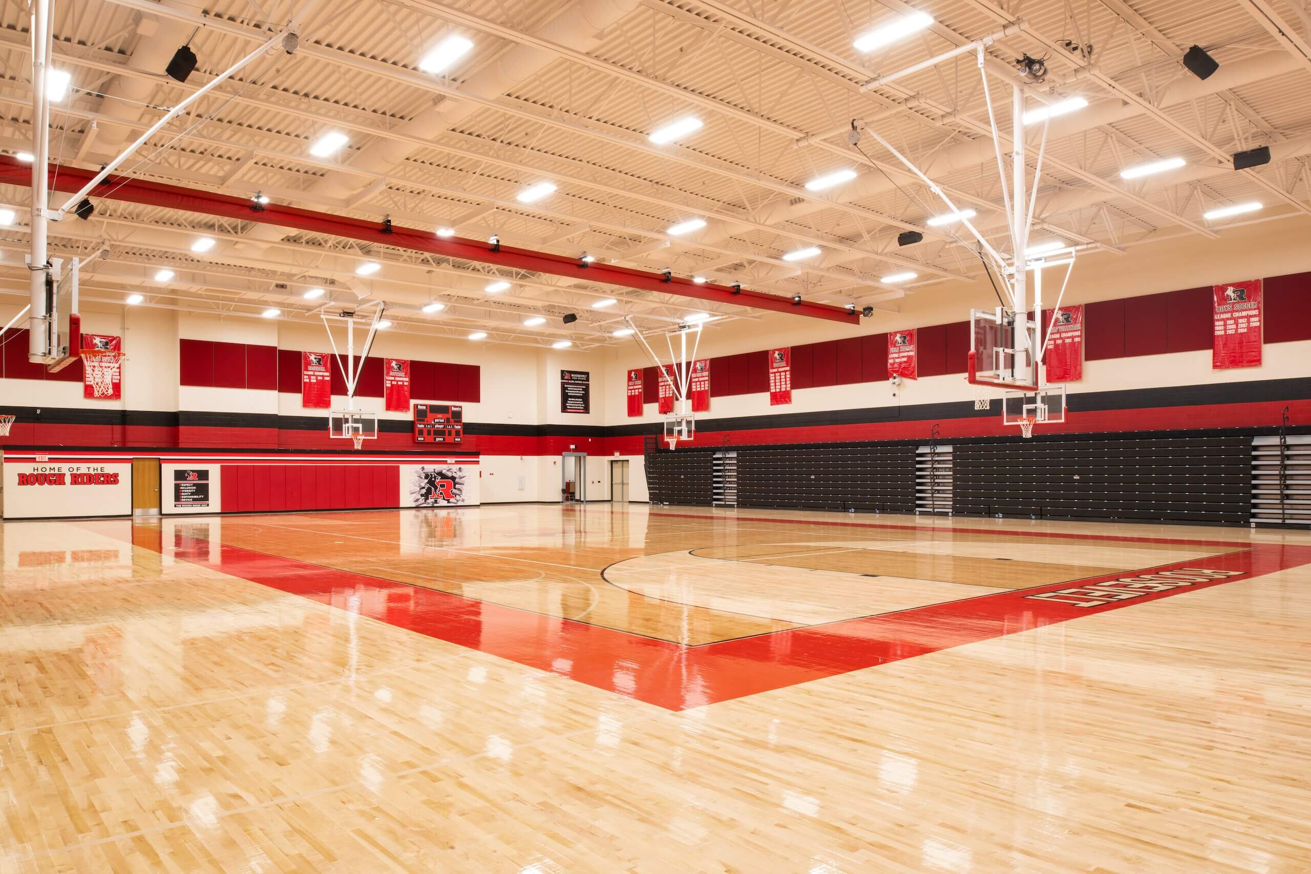 A spacious, empty high school gymnasium with a polished wooden floor, red and black accents, basketball hoops, bleachers, banners, and bright overhead lighting.
