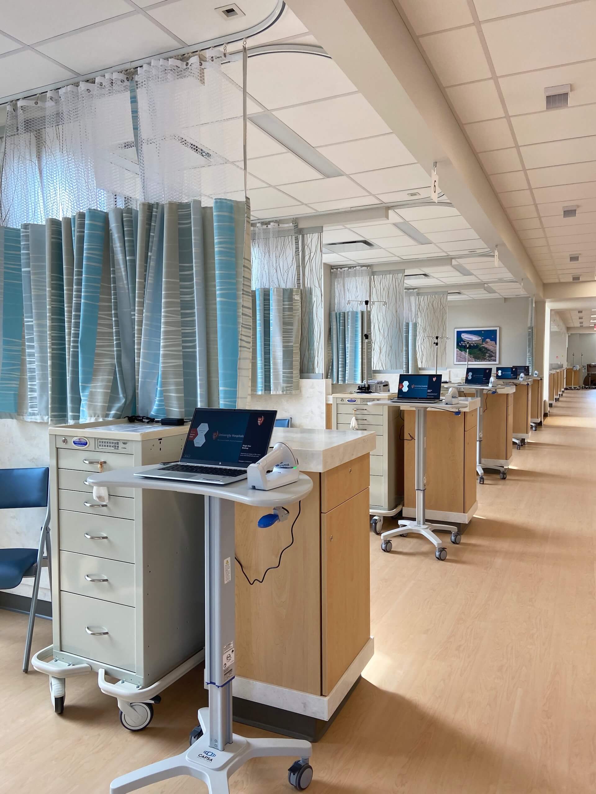 A row of medical carts with laptops lines a hospital ward with blue privacy curtains, wooden cabinets, and bright overhead lighting. The area appears clean and organized.