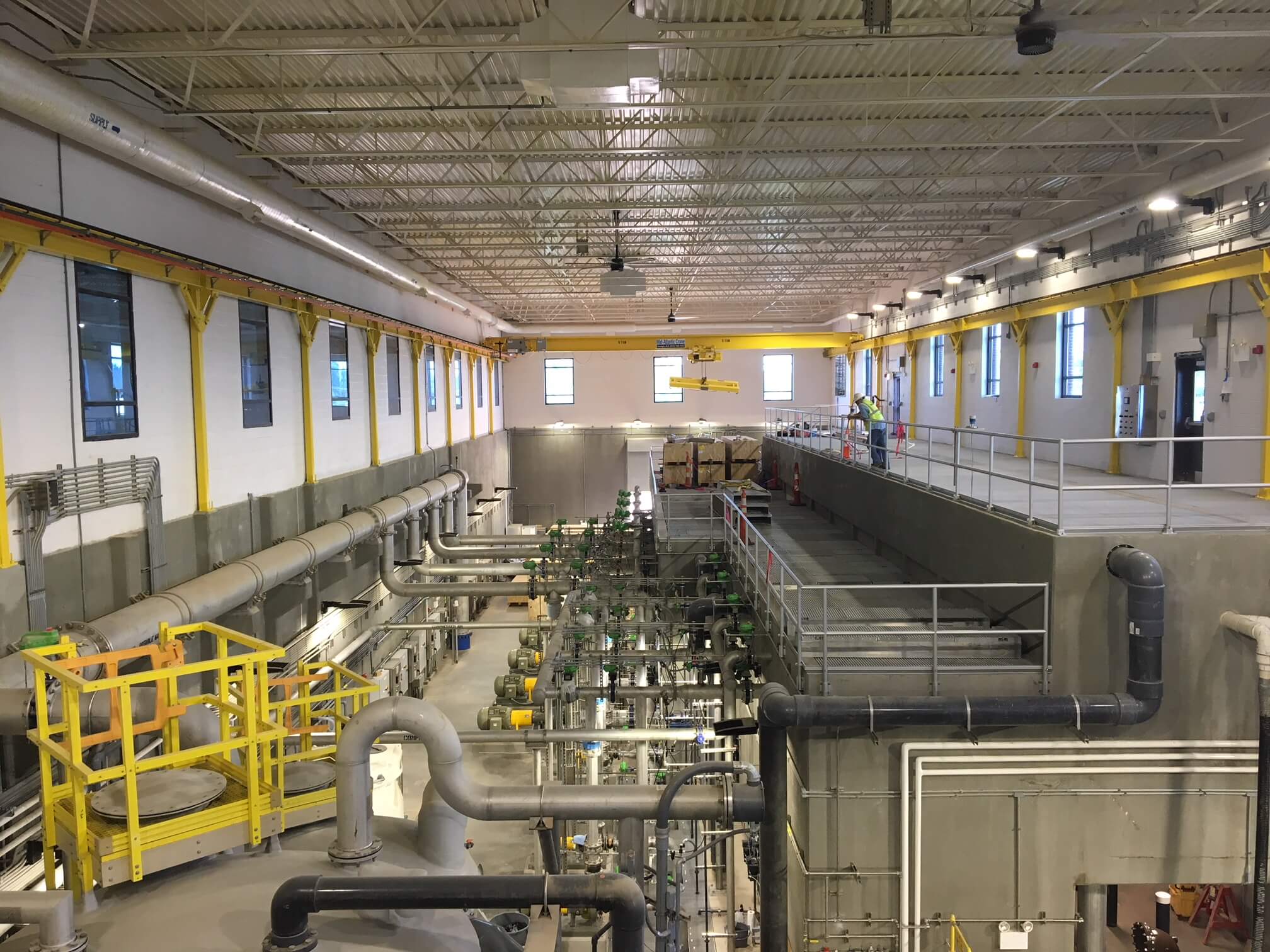 Large, well-lit industrial facility with pipes, platforms, and machinery. Yellow safety rails, high ceilings, and windows line the walls. Several workers are visible on the upper walkway.