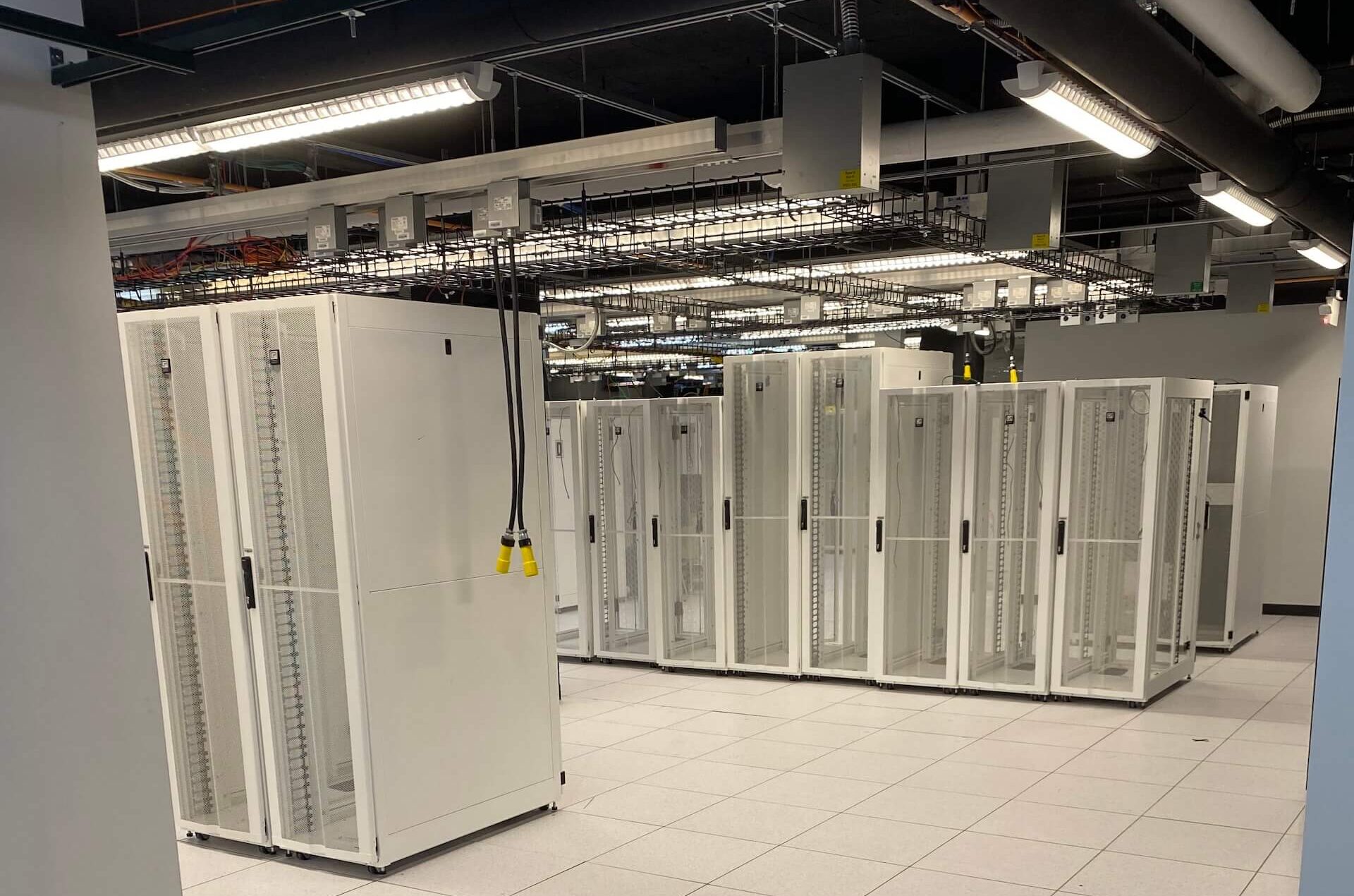 A data center room with multiple empty white server racks on a tiled floor, overhead cable trays, and fluorescent lighting.