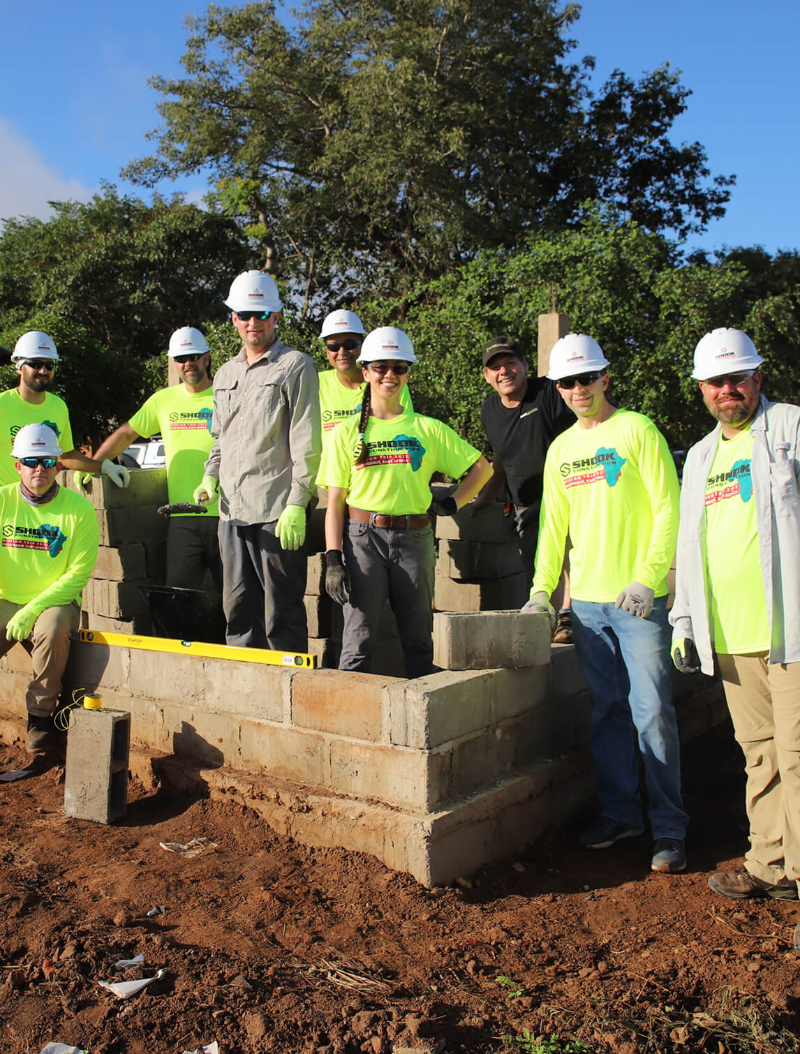 Eight people in hard hats and safety gear pose in front of a partially built brick structure outdoors, surrounded by dirt and trees, with some holding construction tools and smiling at the camera.