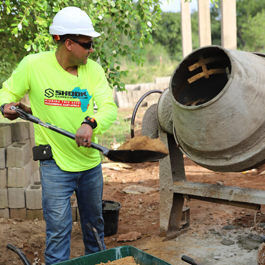 A construction worker in a neon shirt and hard hat shovels sand into a wheelbarrow next to a cement mixer at a building site.