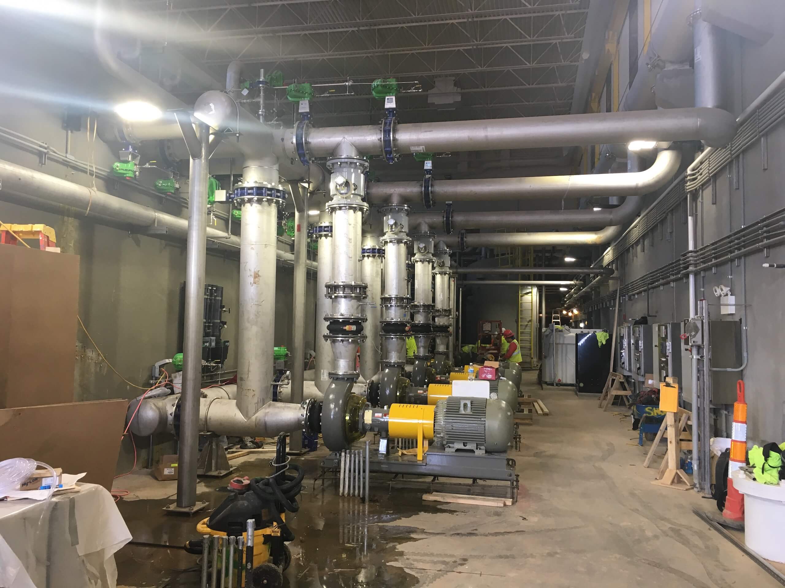 A large industrial room with metal pipes, valves, pumps, and machinery. Workers wearing safety vests are seen in the background. The area has unfinished flooring and equipment scattered around.