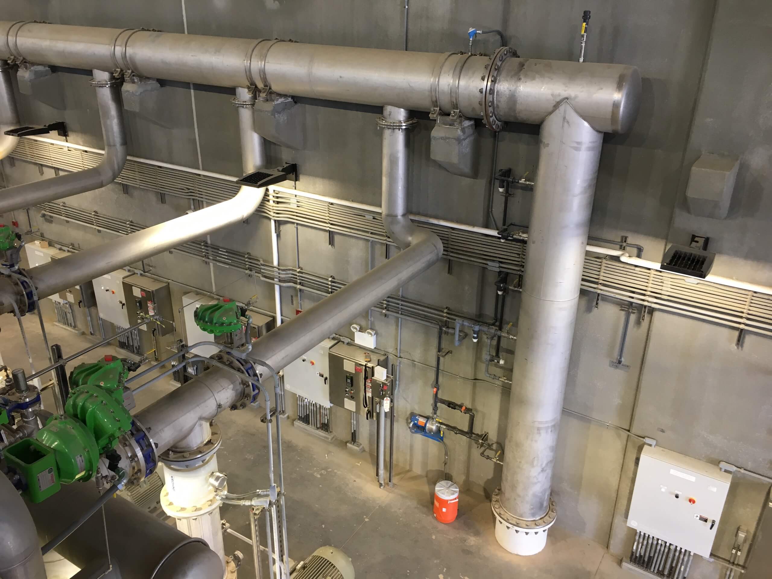 Large metal pipes run along an industrial facility wall with electrical panels, valves, and a red trash bin on the concrete floor below.