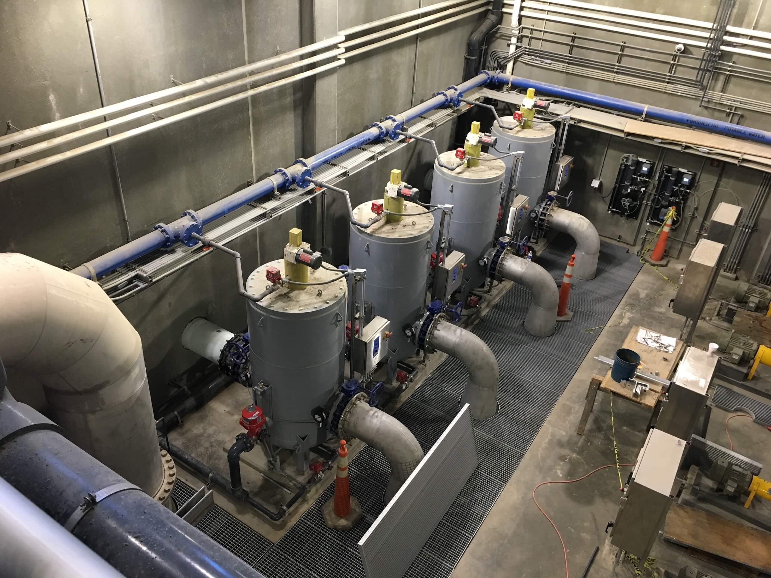 Overhead view of four large cylindrical tanks with pipes, valves, and control panels in an industrial water treatment facility. The area is clean and organized, with safety cones and metal grating on the floor.