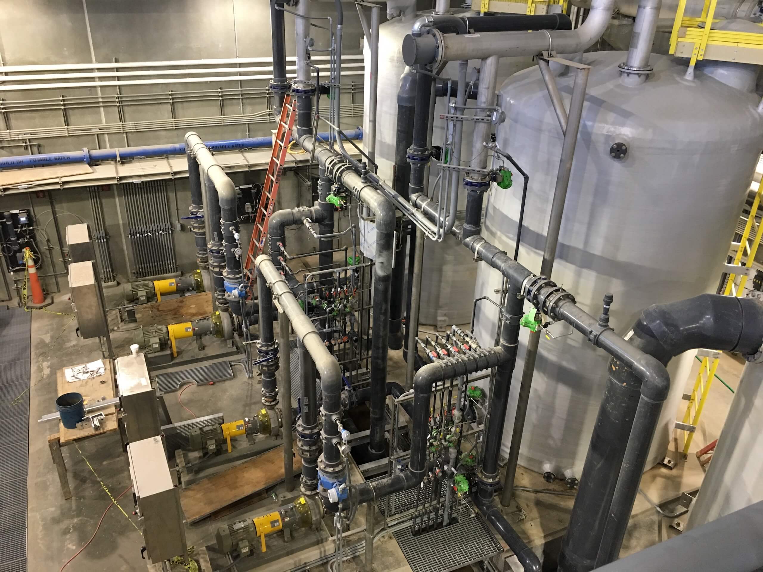 Industrial facility with large tanks, interconnected pipes, valves, control panels, and a red ladder leaning against one tank. The scene is clean, organized, and brightly lit.
