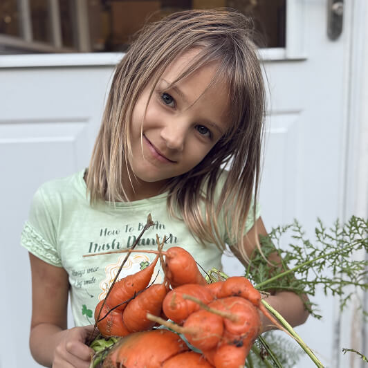 A young girl with light brown hair smiles while holding a bunch of freshly picked, oddly shaped carrots in front of a white door.