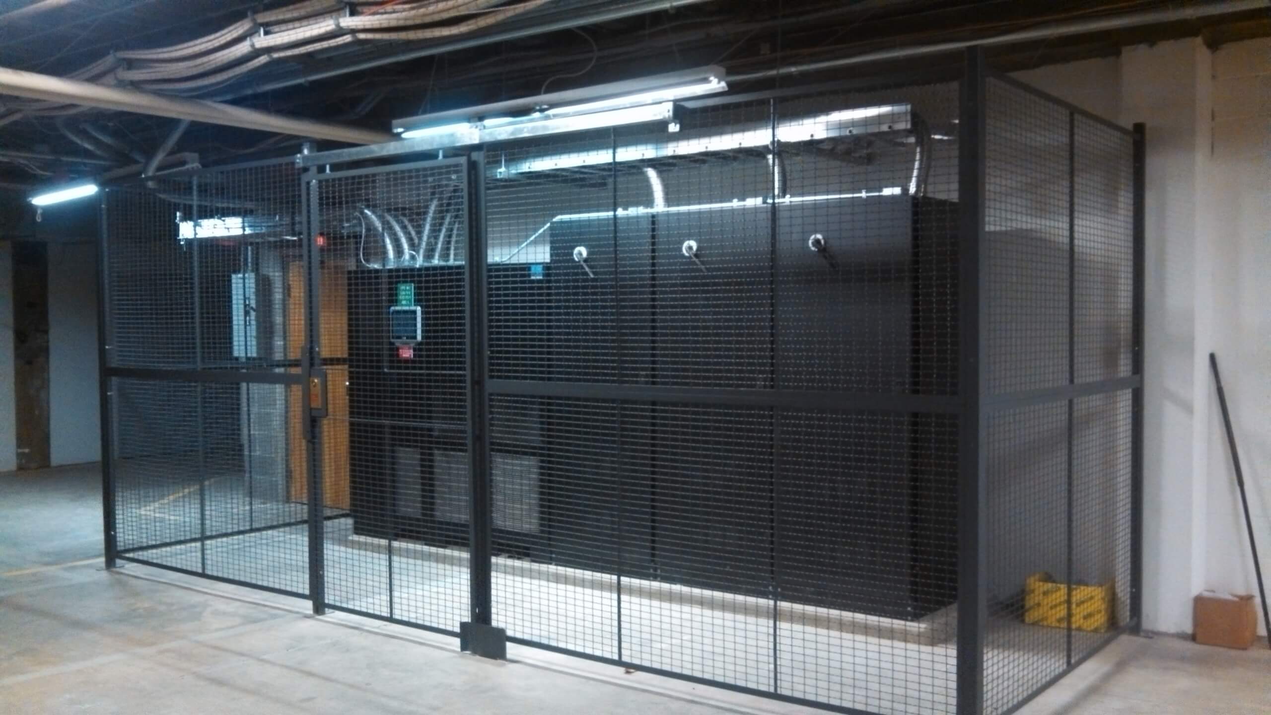 A secure metal cage surrounds electronic equipment and wiring in an industrial or server room, with overhead lights and a concrete floor visible.