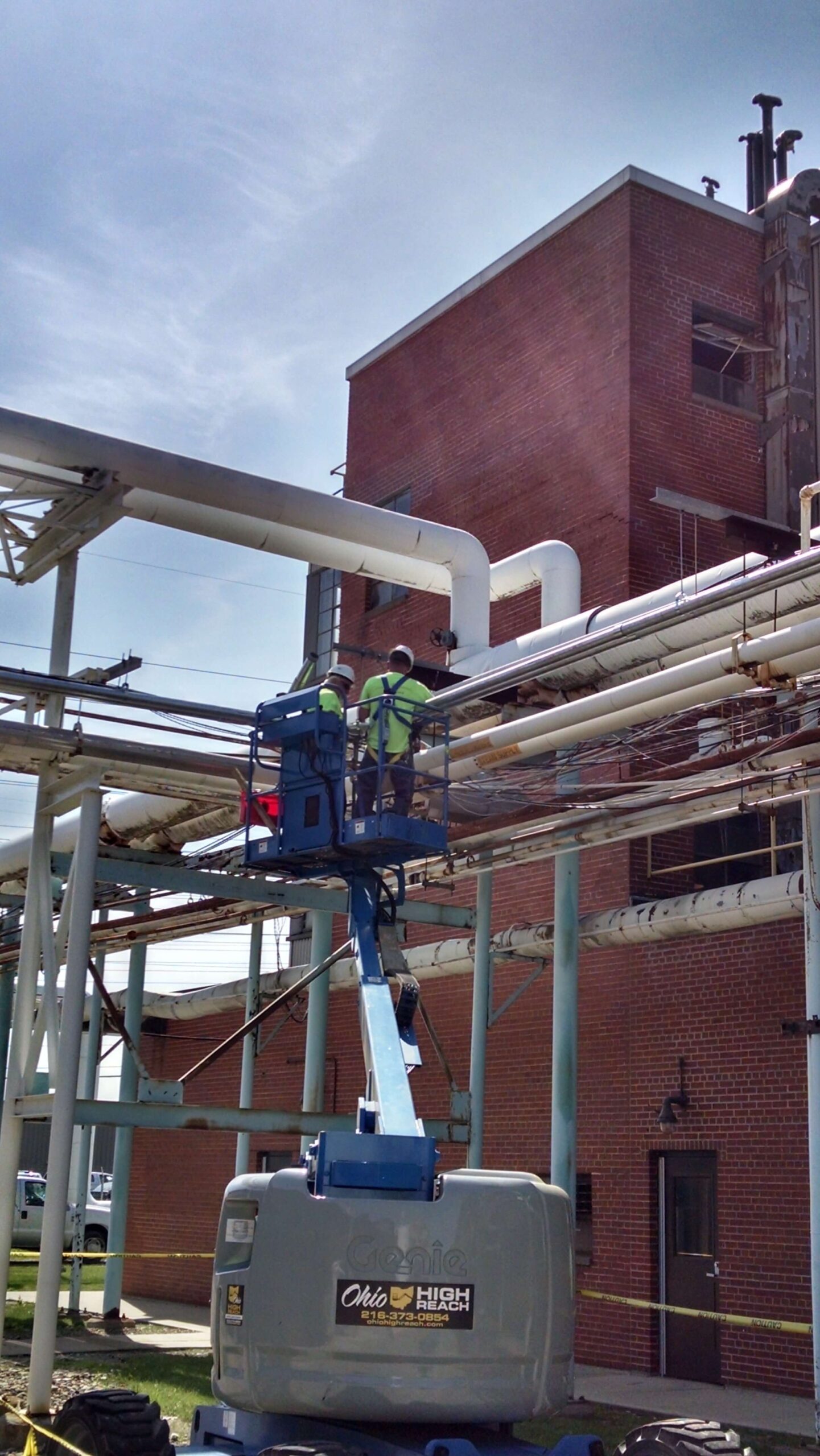 Two workers in safety gear stand on a blue lift inspecting or repairing pipes outside a brick industrial building on a sunny day.