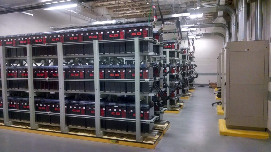 Rows of large industrial batteries are organized on metal racks inside a clean, well-lit facility with concrete floors and exposed ceiling pipes.