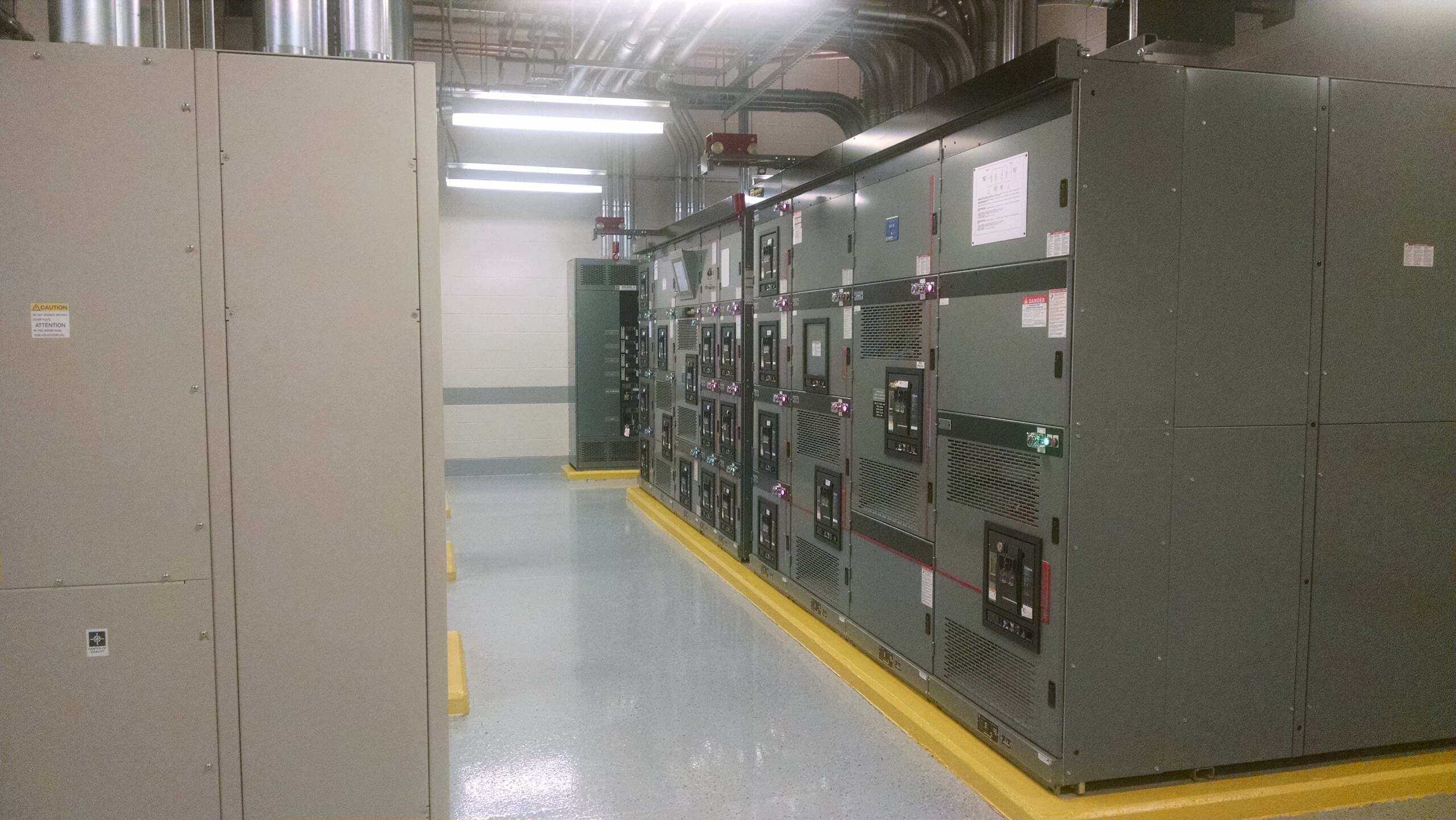 A clean industrial room with large electrical control panels and metal cabinets, bright overhead lighting, and visible pipes on the ceiling. The floor is polished and reflective, with safety yellow borders.