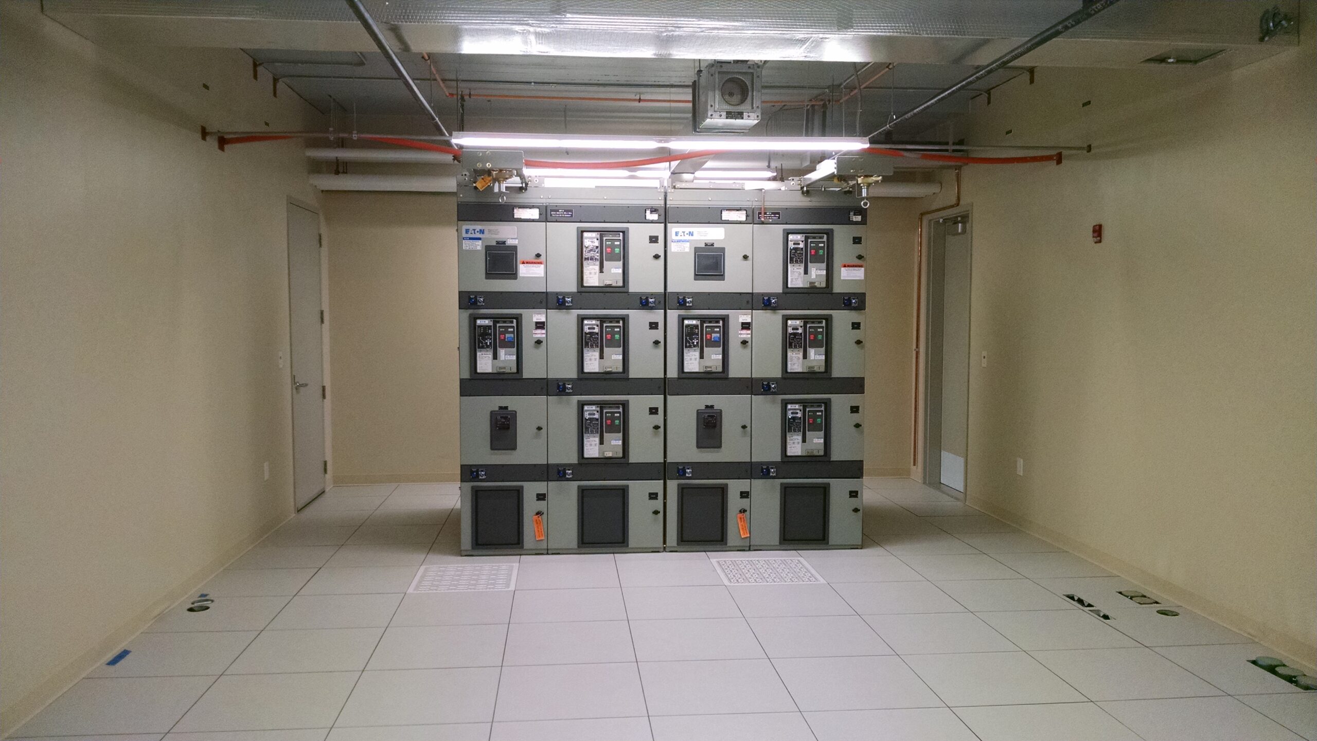 A control room with light walls and floor featuring a large electrical panel with switches and meters; two doors are visible on either side of the room.