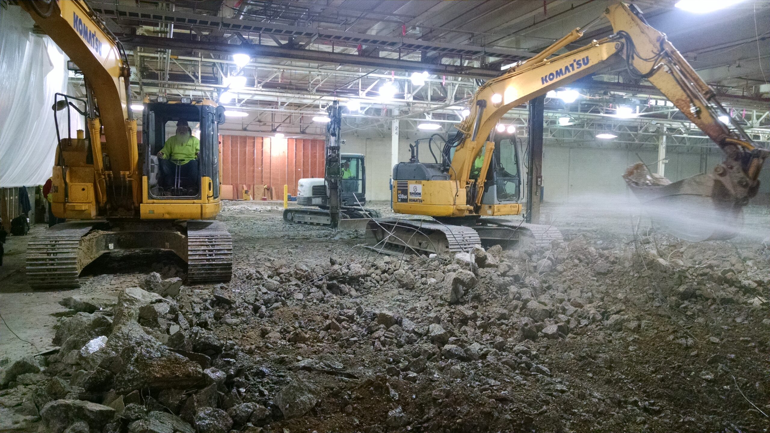 Two yellow Komatsu excavators break up concrete on the floor inside an industrial building, with debris and rubble scattered around. Bright overhead lights illuminate the construction area.