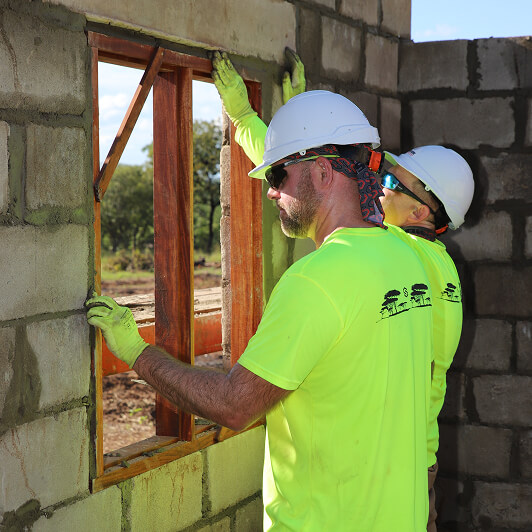 Two construction workers in bright yellow shirts and white helmets install a wooden window frame into a cinder block wall of an unfinished building on a sunny day.
