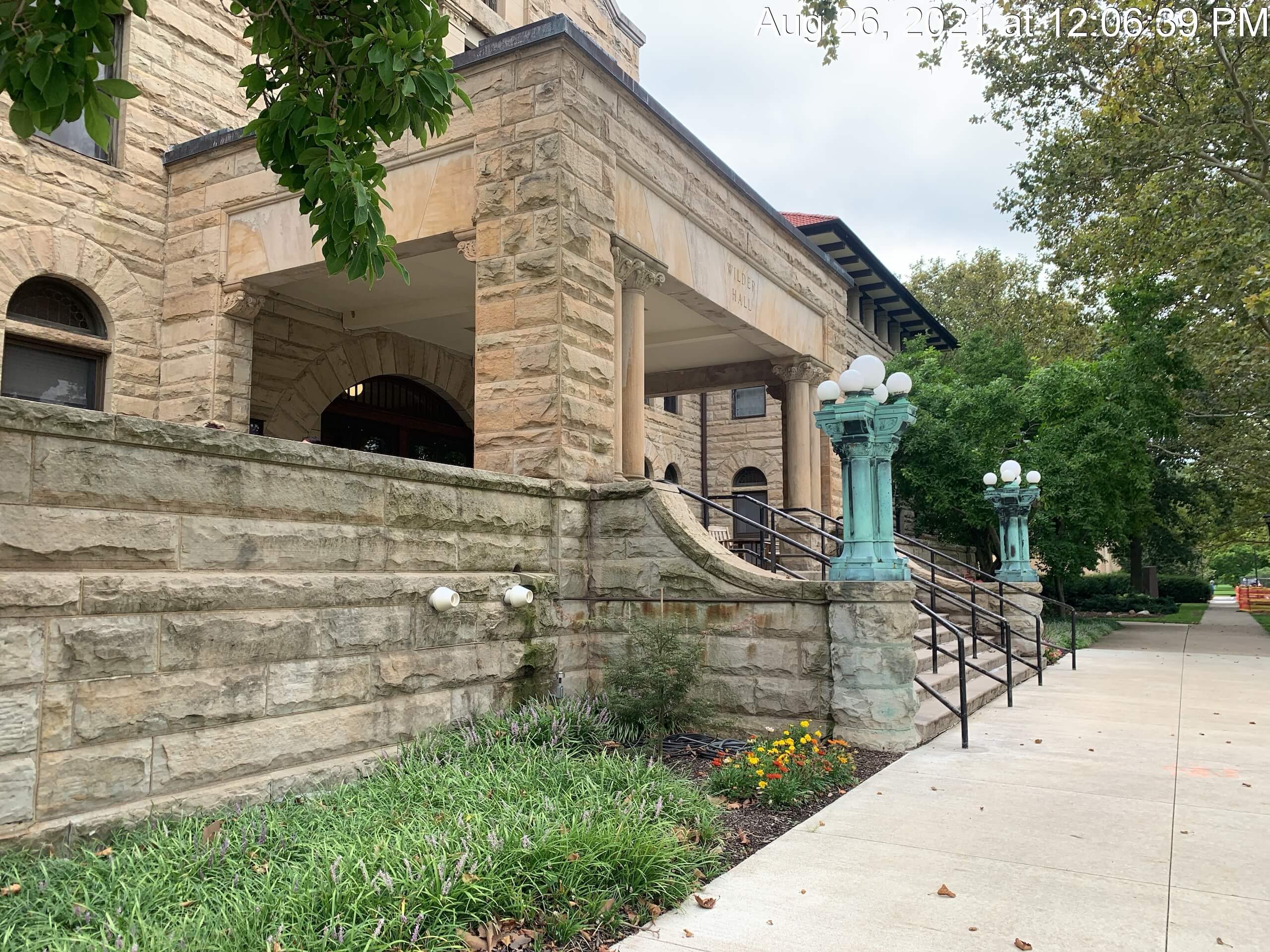 A historic stone building with arched windows, a ramp with green lamp posts, and a garden with flowers along a sidewalk. The date and time are visible in the top-right corner.