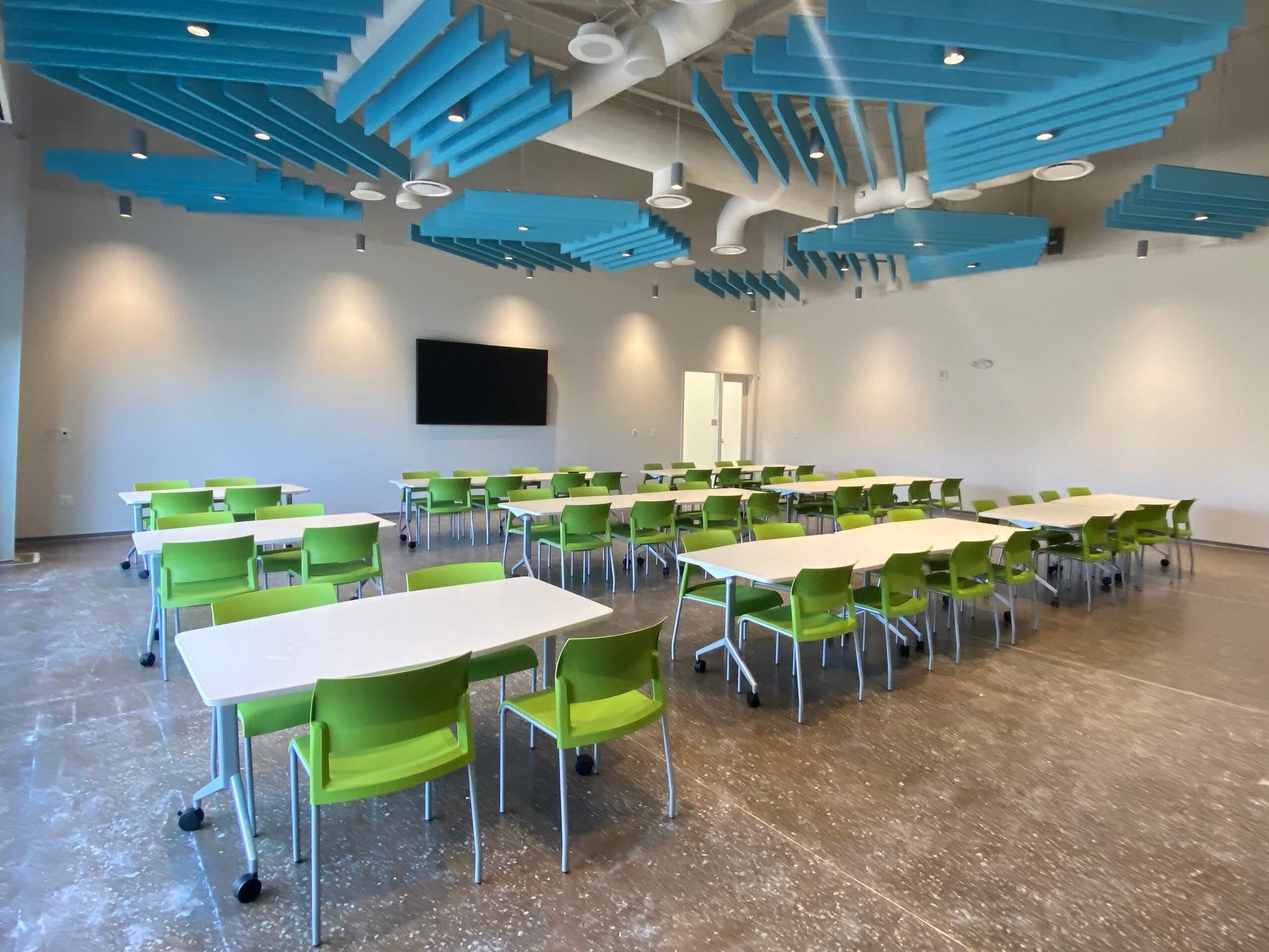 A modern classroom with white tables, green chairs, a wall-mounted screen, and blue acoustic panels on the ceiling. The room is well-lit and spacious, with a concrete floor and minimal decor.