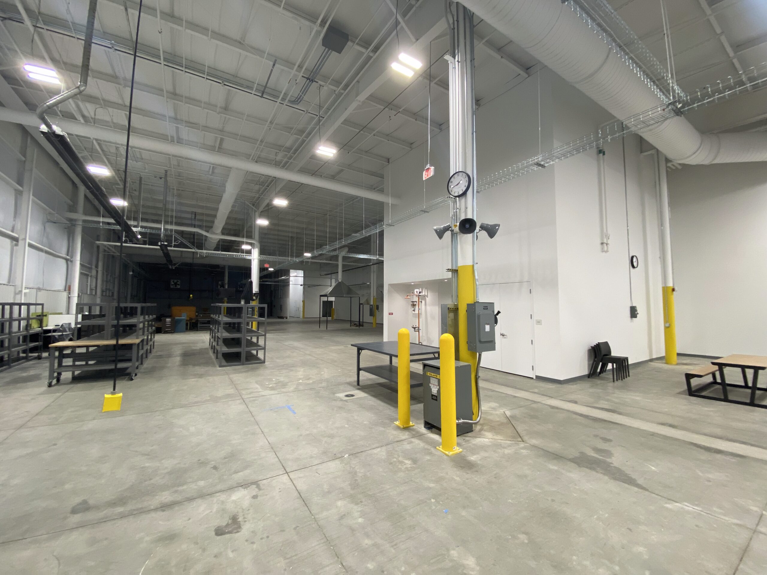 A spacious industrial warehouse with high ceilings, empty metal shelving, tables, and yellow safety barriers. The area is well-lit with overhead lights and has a polished concrete floor.