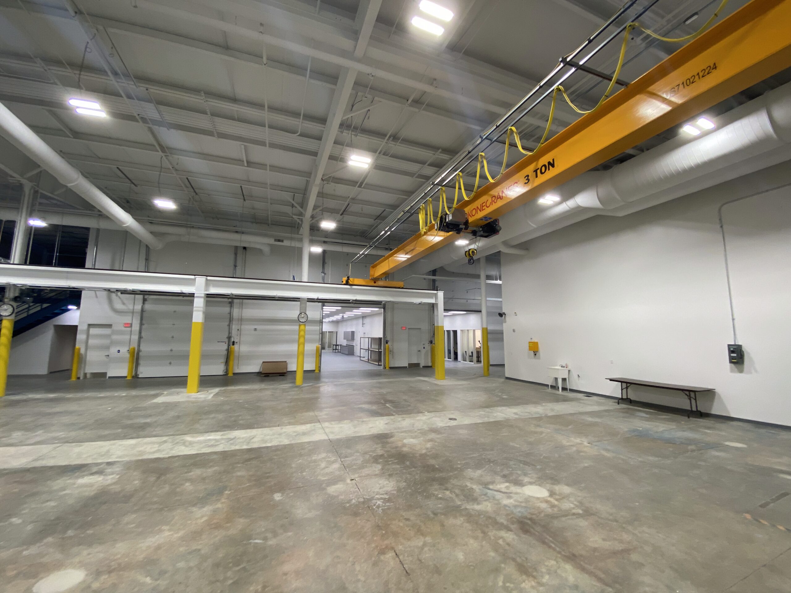 Spacious industrial warehouse with high ceilings, yellow support columns, a large overhead crane labeled 3 TON, and a few tables along the walls. The floor is concrete and the area is well-lit.