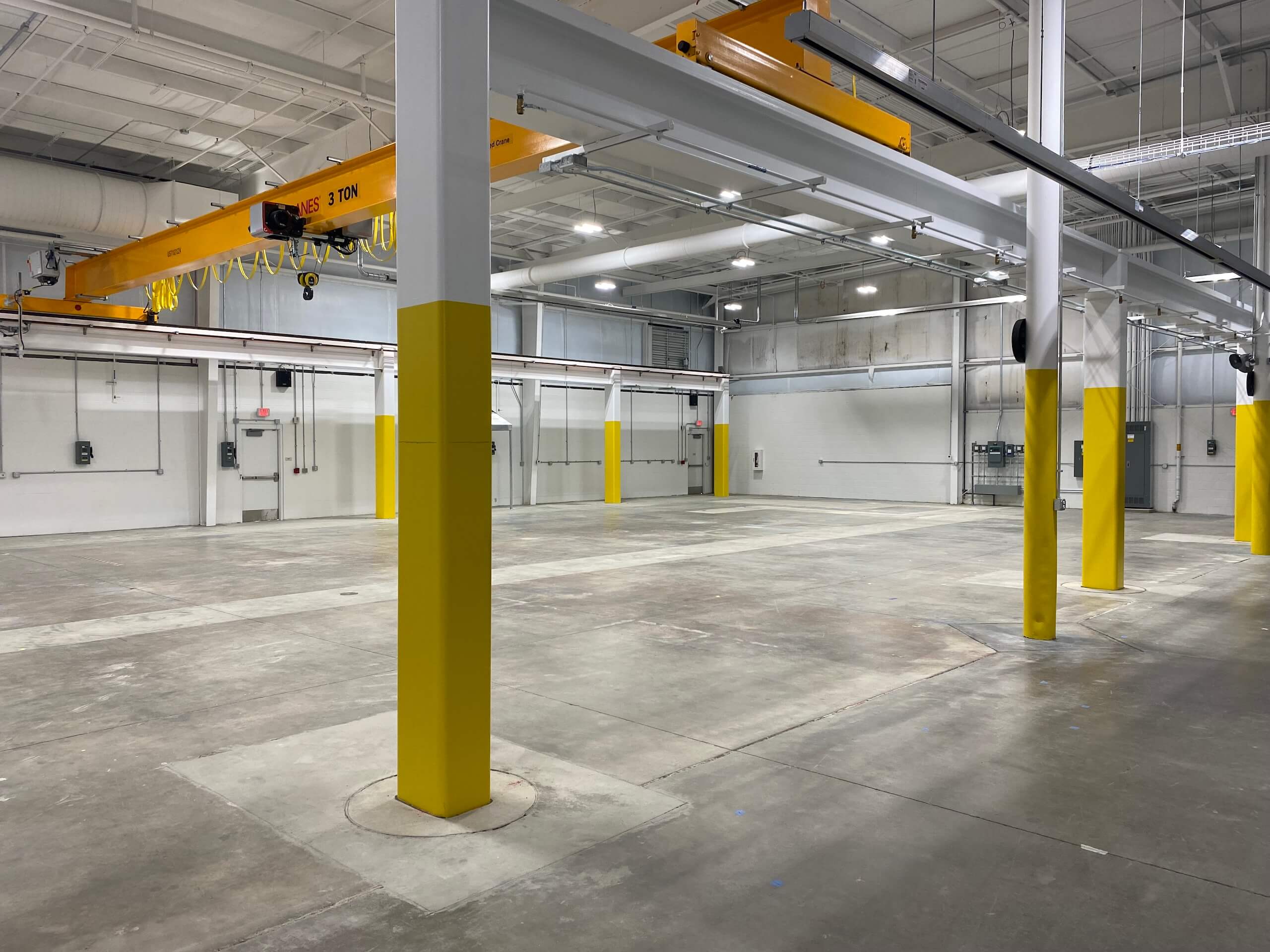 Spacious empty warehouse with yellow pillars, overhead crane system, concrete floor, and white walls. Bright lighting fills the industrial interior.