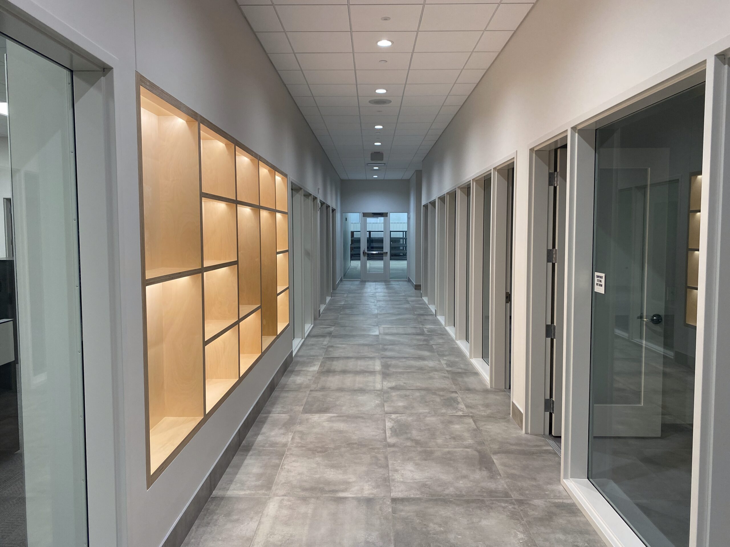 Modern hallway with gray tile floor, glass office doors on the right, and built-in wooden display shelves on the left, leading to a glass-walled area at the end of the corridor.