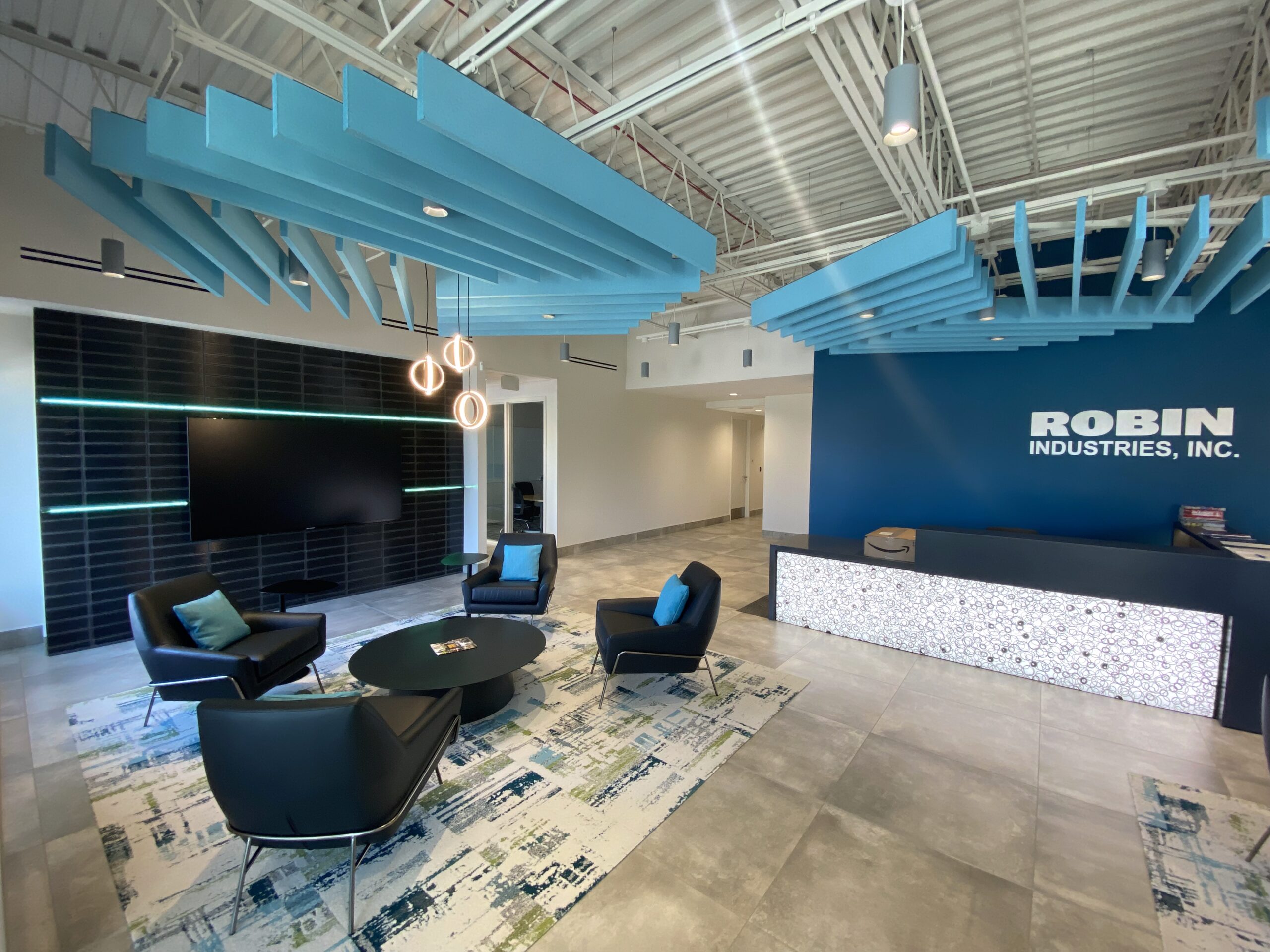 Modern office lobby with blue ceiling panels, black chairs around a coffee table, a TV on the wall, decorative lights, a reception desk, and a blue accent wall with ROBIN INDUSTRIES, INC. written on it.