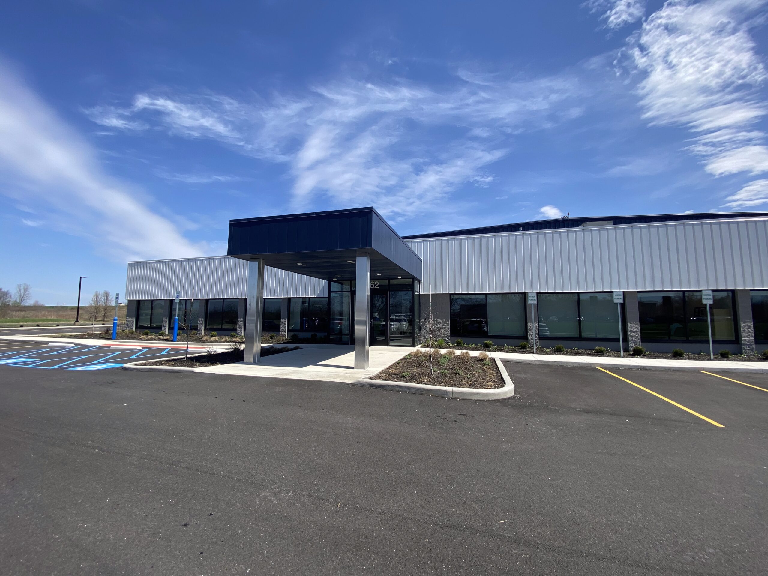 Modern, single-story commercial building with metal siding, large glass windows, and a covered entrance. The parking lot includes handicap spaces. The sky is blue with wispy clouds.