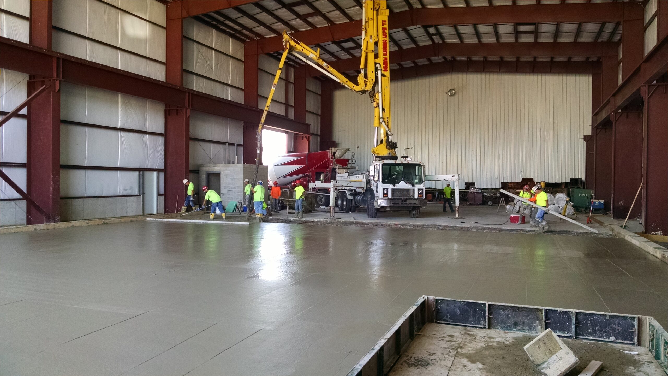 A group of construction workers in safety gear pour and level fresh concrete inside a large metal-framed industrial building using a concrete pump truck.