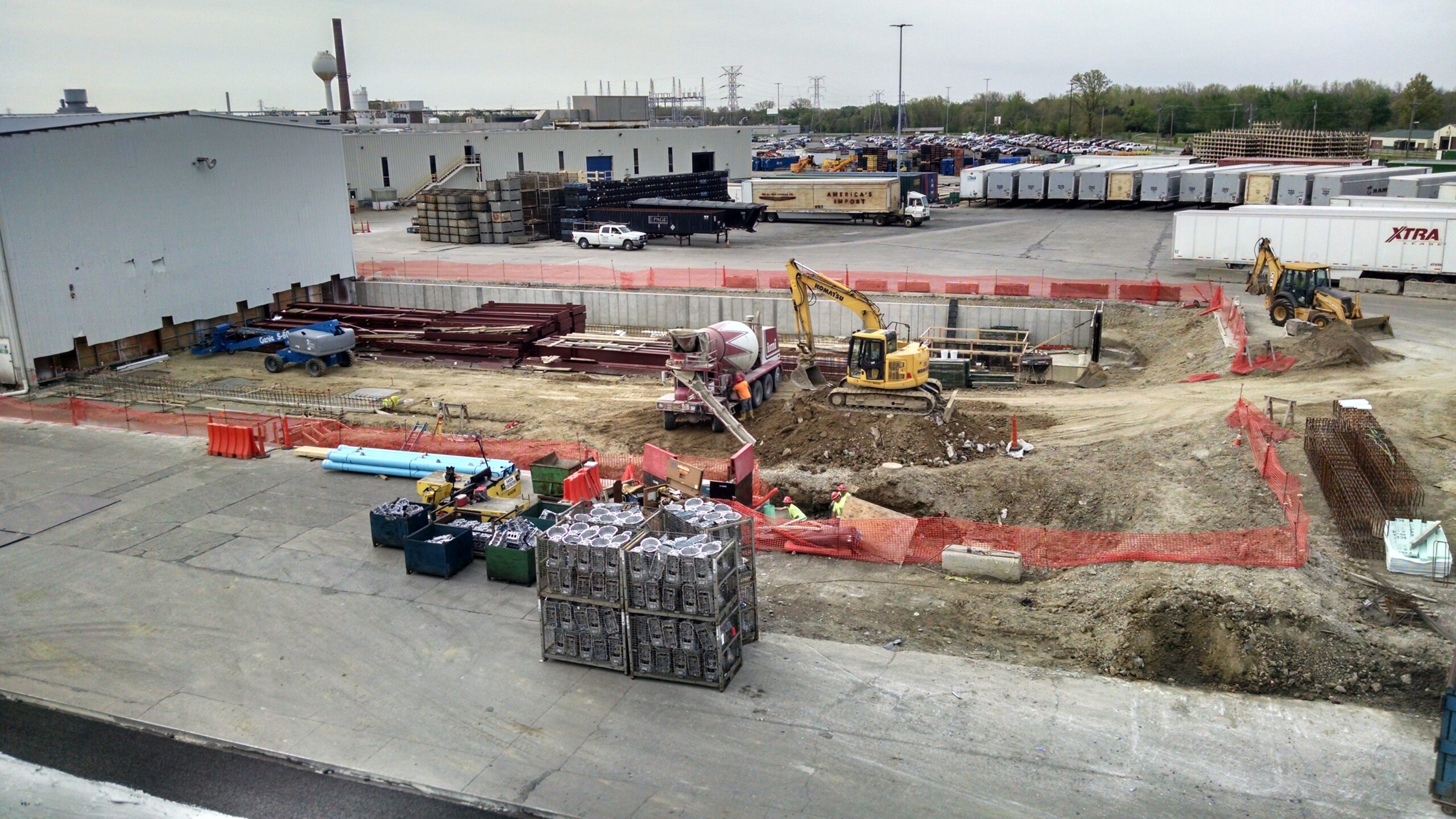 A construction site with heavy machinery, workers, concrete mixer, steel beams, and various building materials surrounded by orange safety fencing and industrial buildings in the background.