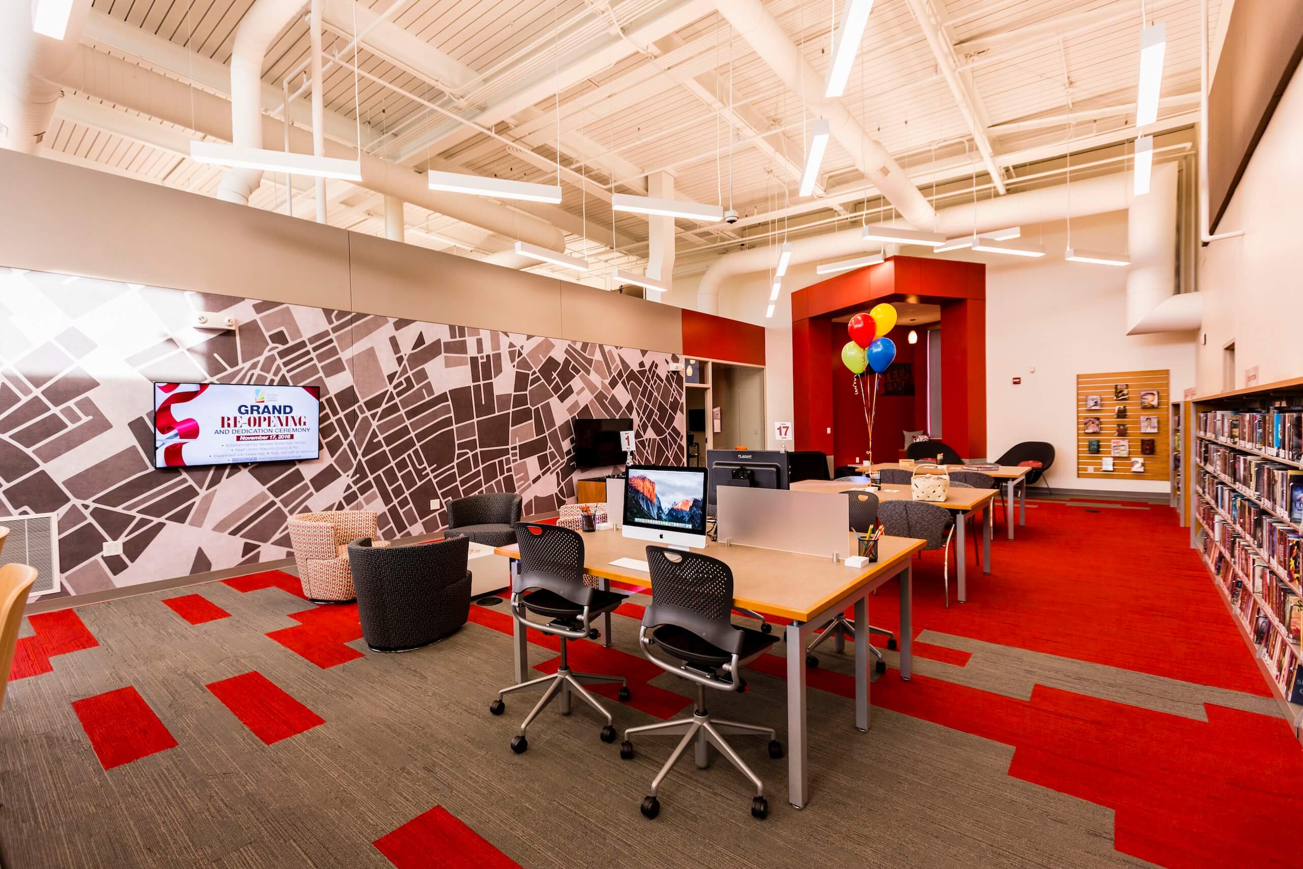 Modern library interior with computers on desks, bookshelves on the right, a wall-mounted TV screen on the left, red and gray carpeting, and balloons in a corner by an alcove with chairs.
