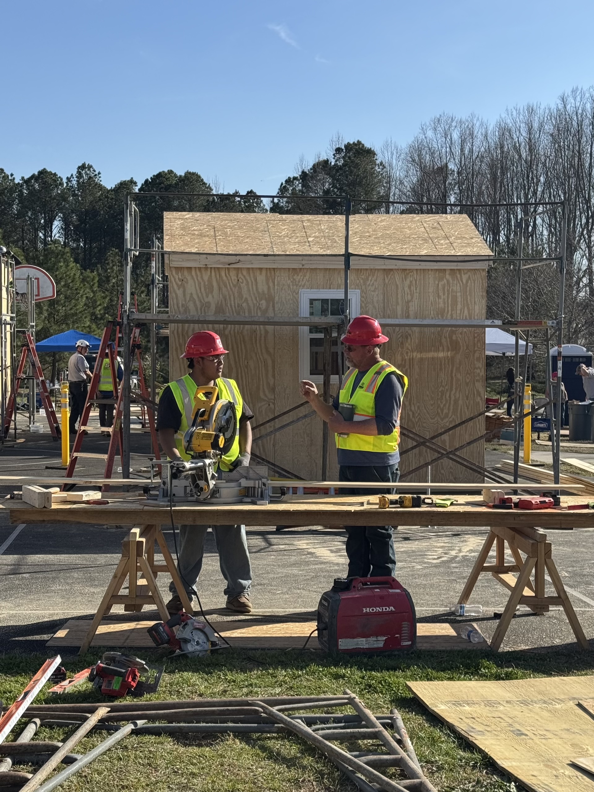 Two construction workers in red helmets and yellow vests talk near a saw and tools, with a wooden structure being built behind them on a sunny day.