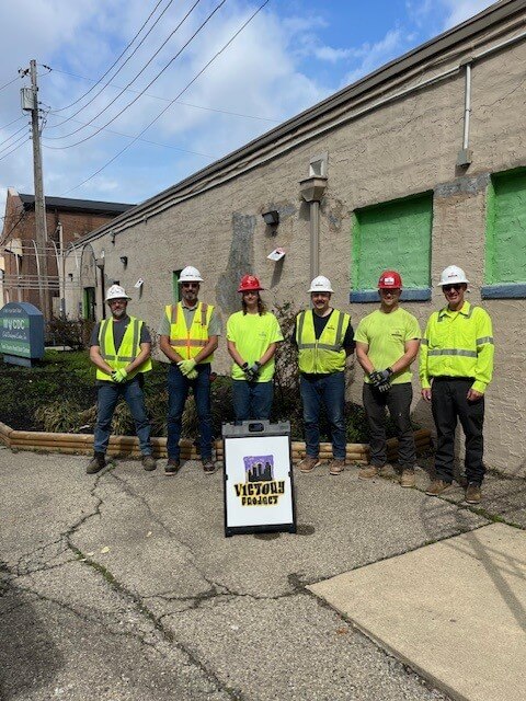 Six construction workers in safety gear stand outside a tan building with green window panels, behind a sign that reads Victory Projects. The sky is partly cloudy and theres some greenery nearby.