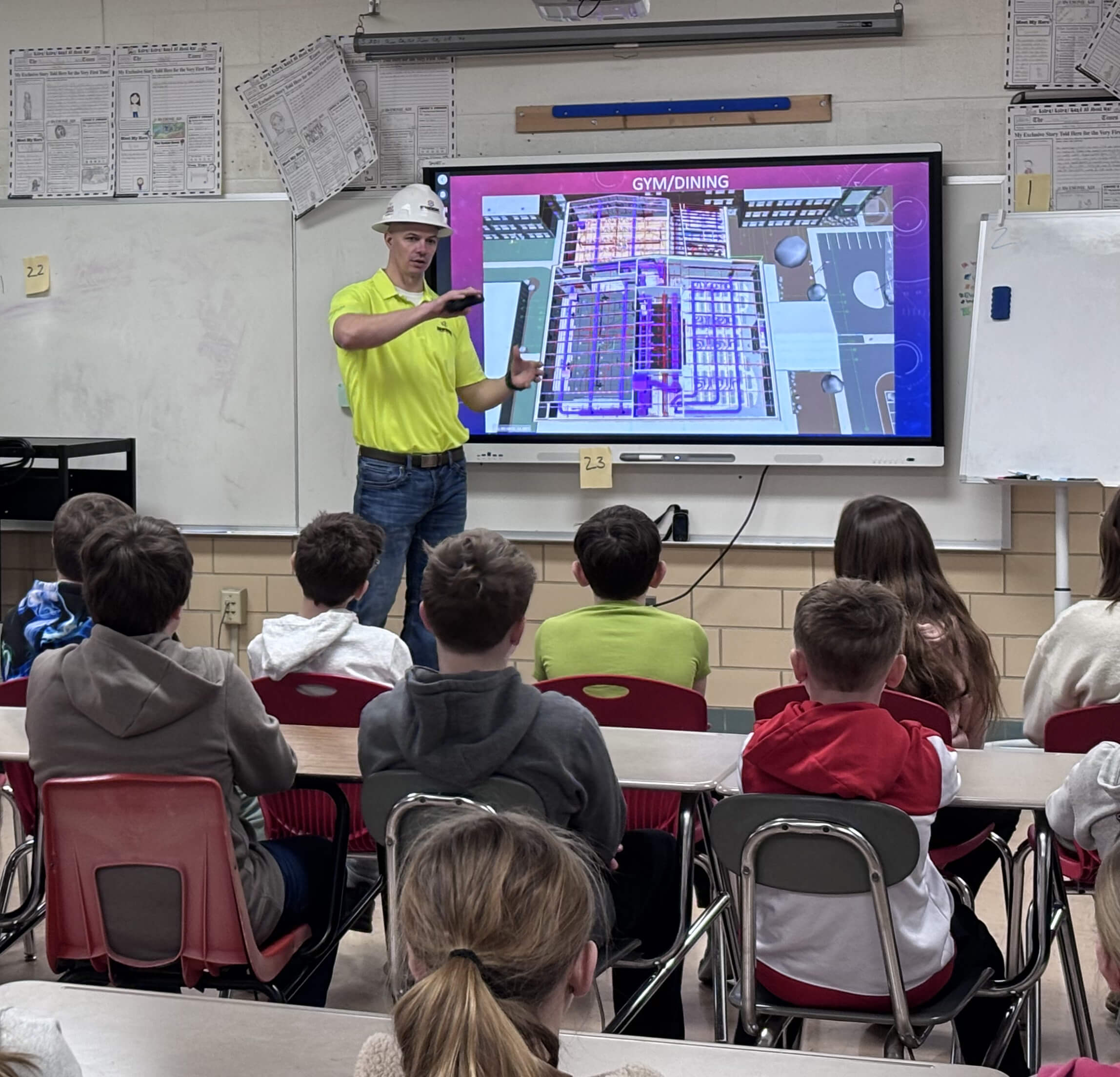 A man in a yellow shirt and hard hat speaks to a classroom of children while pointing at a large screen displaying a digital building layout. The students are seated and facing him attentively.