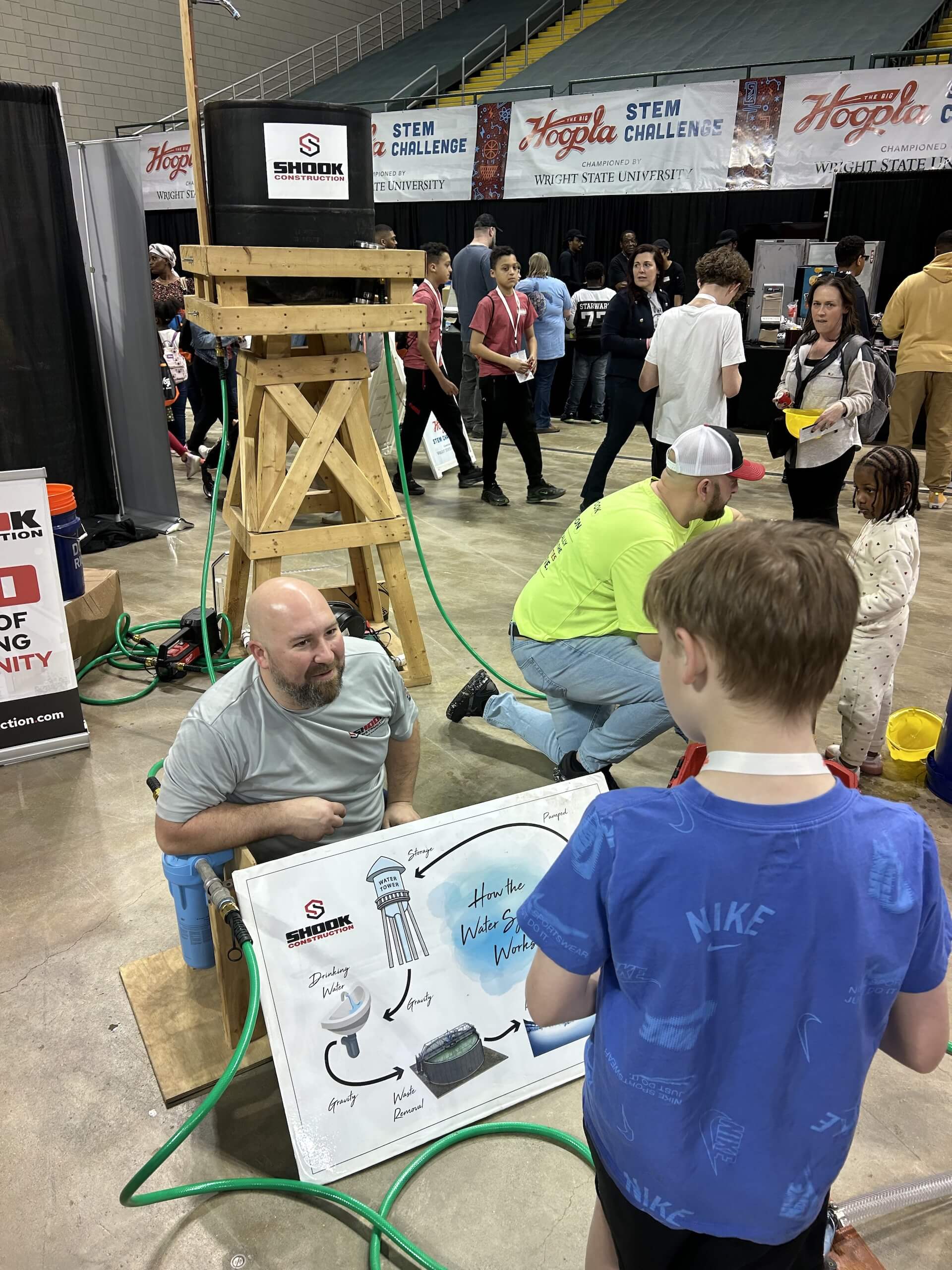 A man talks to a boy at a STEM event booth with a wooden structure, hoses, and a water system diagram. People walk and interact in the busy event space.