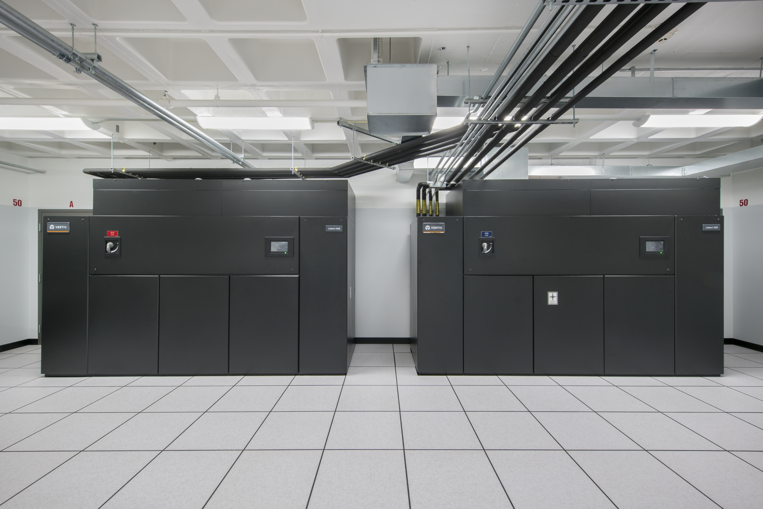 Two large black server or data center units stand side by side in a white room with tiled floors and ceiling pipes. Each unit has a control panel and indicator lights.